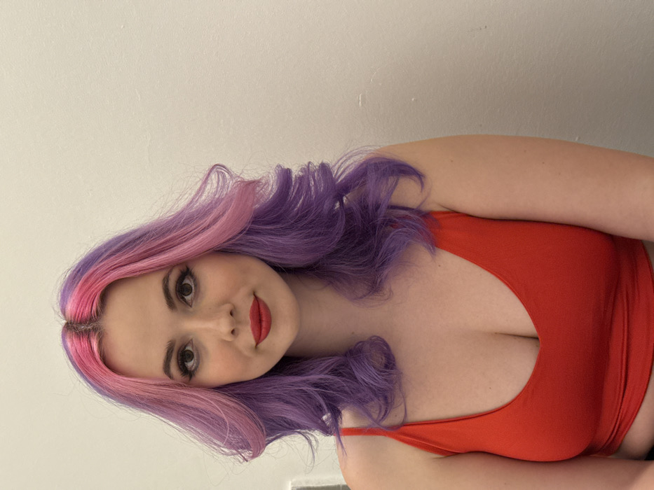 A fair-skinned woman with shoulder-length, wavy pink and purple hair is looking at the camera. She is wearing a red tank top and red lipstick. The background is a light, neutral color. The lighting is bright and even.