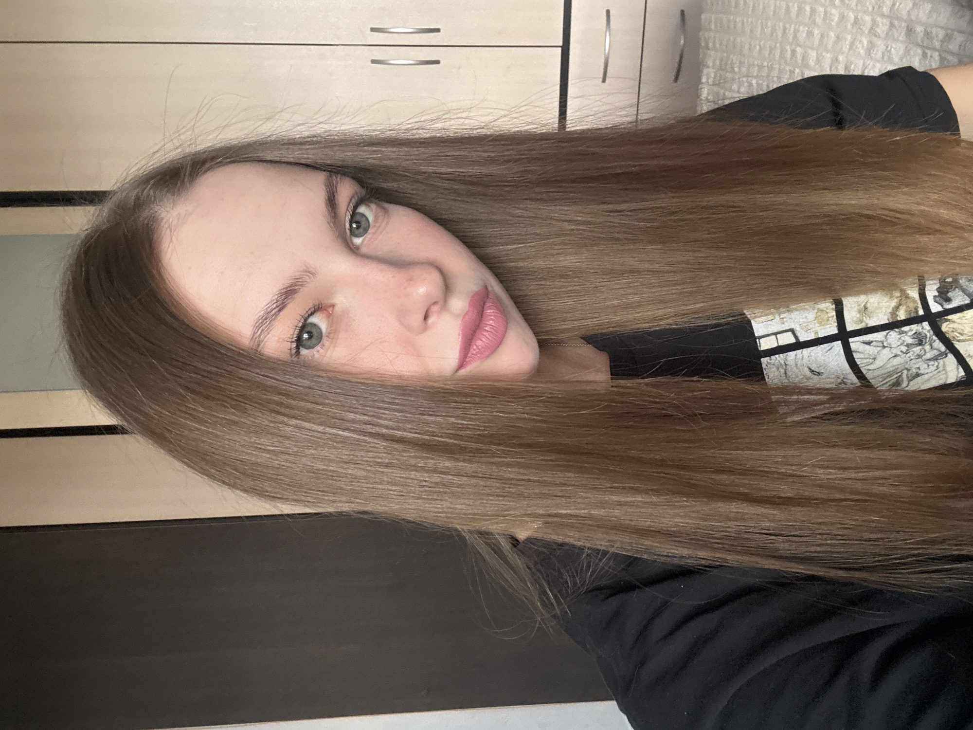 A young woman with long, straight, brown hair and green eyes is shown from the chest up, looking directly at the camera. She is wearing a black t-shirt with a graphic print. Her lips are painted a light pink. She has freckles on her nose and cheeks. The background appears to be an indoor setting, possibly a bedroom, with a dark wardrobe on the left and a light-colored cabinet with handles on the right. The image quality is clear. There is no information to suggest the location is the city of Perm, Russia.