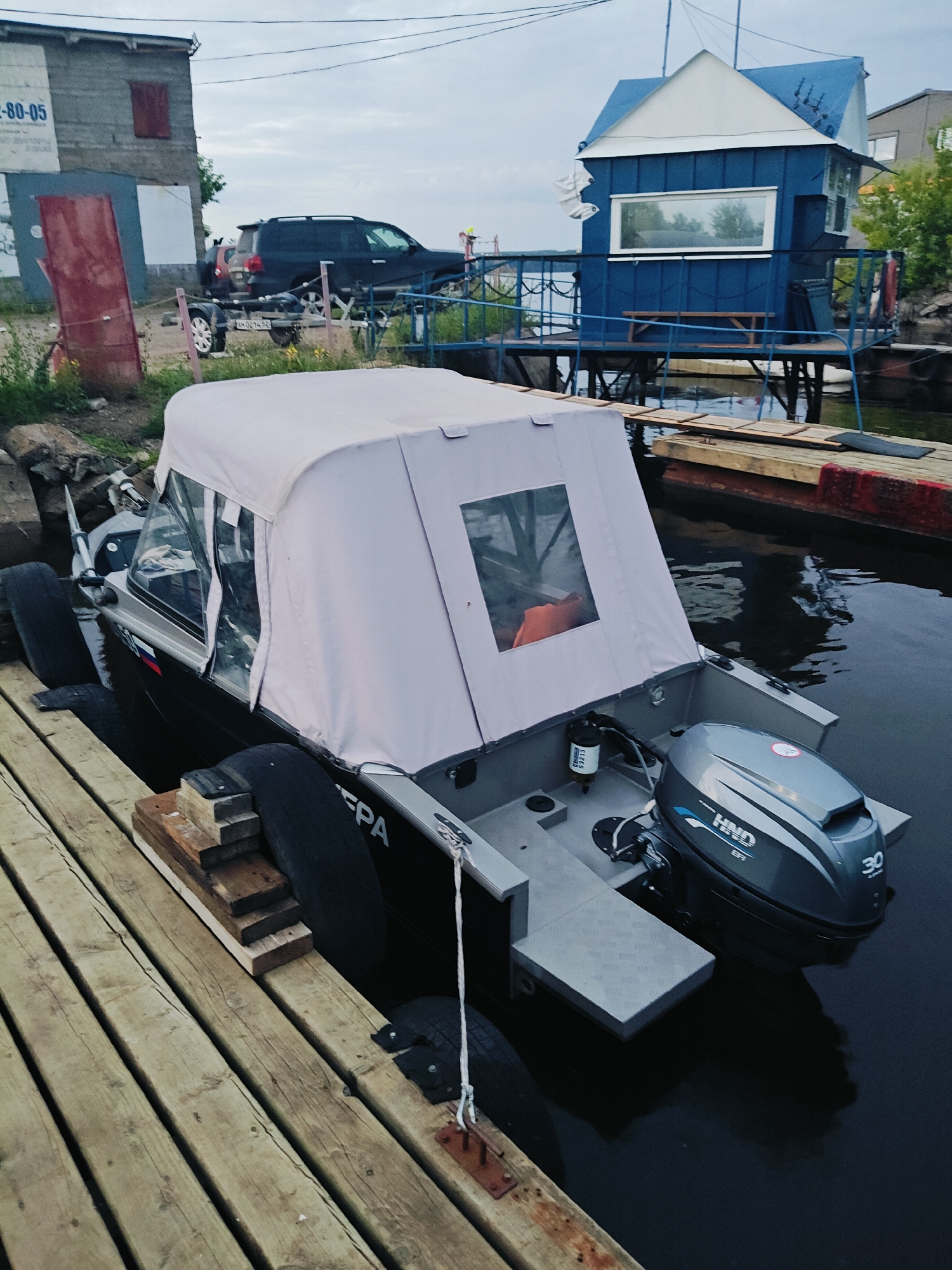 A wide shot captures a boat docked at a wooden pier on a cloudy day in Chaikovsky, Russia. The boat, a dark grey vessel with a light grey canopy and windows, is secured by thick black tires acting as fenders. An outboard motor, branded "HND" with "30" visible, is attached to the stern. The pier, constructed from weathered wooden planks, extends from the lower left of the frame. In the background, a small, blue, multi-story building with a white roof stands on stilts over the water. To the left of the frame, a grey industrial building with a red door is visible, along with a dark-colored SUV and a trailer. The water is dark and still, reflecting the overcast sky.