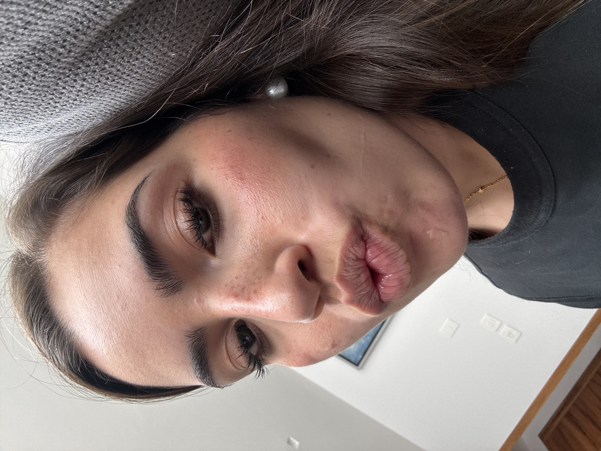 A close-up, slightly angled, selfie of a woman with dark hair and light skin. Her lips are puckered, suggesting a kiss. She has defined eyebrows, dark eyelashes, and freckles on her nose. She wears a black t-shirt and a delicate gold necklace. A pearl earring is visible. The background shows a gray textured surface near her hair, a white wall, a glimpse of a framed picture, and a portion of a wooden frame. There is no indication of San Bernardino Tlaxcalancingo, Mexico, in the image.