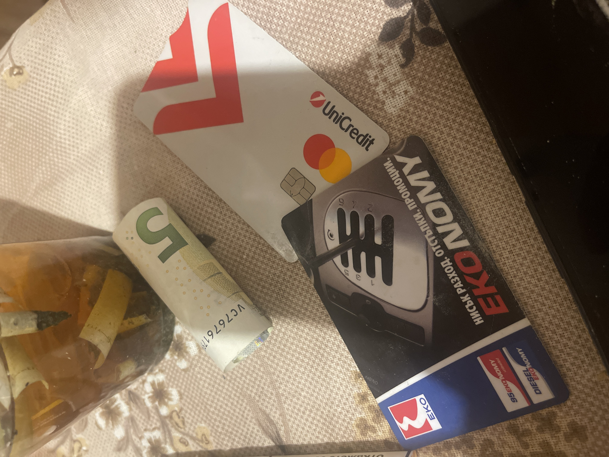 An overhead, slightly tilted shot captures several everyday items resting on a patterned, light-colored fabric surface, possibly a tablecloth. The primary subjects are three distinct objects: a bank card, a loyalty card, and a rolled-up banknote, alongside a container with cigarette waste.
In the upper-middle of the frame is a white UniCredit bank card, featuring the bank's red "U" logo and "UniCredit" text. Below the bank name, a classic Mastercard logo with overlapping red and orange circles is visible, along with the metallic EMV chip. Partially obscured beneath it and extending to the right is a dark-colored loyalty card for EKO. This card displays a prominent graphic of a manual gear shifter with gear numbers (1, 3, 5, 2, 4, 6) and the brand "EKO" in white text against a red square. It also features the word "EKONOMY" and slogans in Cyrillic "НИСЪК РАЗХОД. ОТСТЪПКИ. ПРОМОЦИИ." (LOW EXPENSE. DISCOUNTS. PROMOTIONS.), as well as "95EKONOMY unleaded" and "DIESEL EKONOMY".
To the left, a green 5 Euro banknote is rolled up, partially revealing its face value "5" and the serial number "VC7676179". Next to the banknote, on the far left, a transparent glass or plastic container is visible, filled with numerous used cigarette butts, some with ash and filters clearly discernible.
The overall scene suggests a casual indoor setting, with no specific activity taking place beyond the arrangement of these items. The lighting appears to be ambient indoor light.