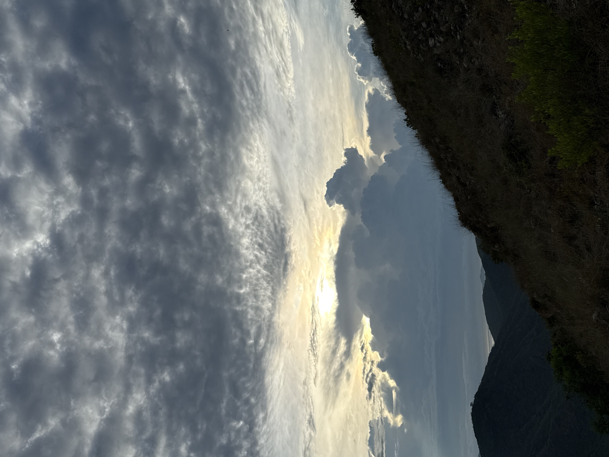 The image shows a view from a high vantage point in or near Bac Nha Trang Ward, Vietnam. The sky is filled with a mix of altocumulus and cirrus clouds, partially obscuring the sun. The light from the sun creates a bright area in the clouds. To the right of the sky, there is a steep, rocky slope covered with sparse vegetation, including some patches of green grass. In the distance, there are mountain peaks visible, suggesting the landscape is mountainous. The overall impression is one of natural scenery and altitude.