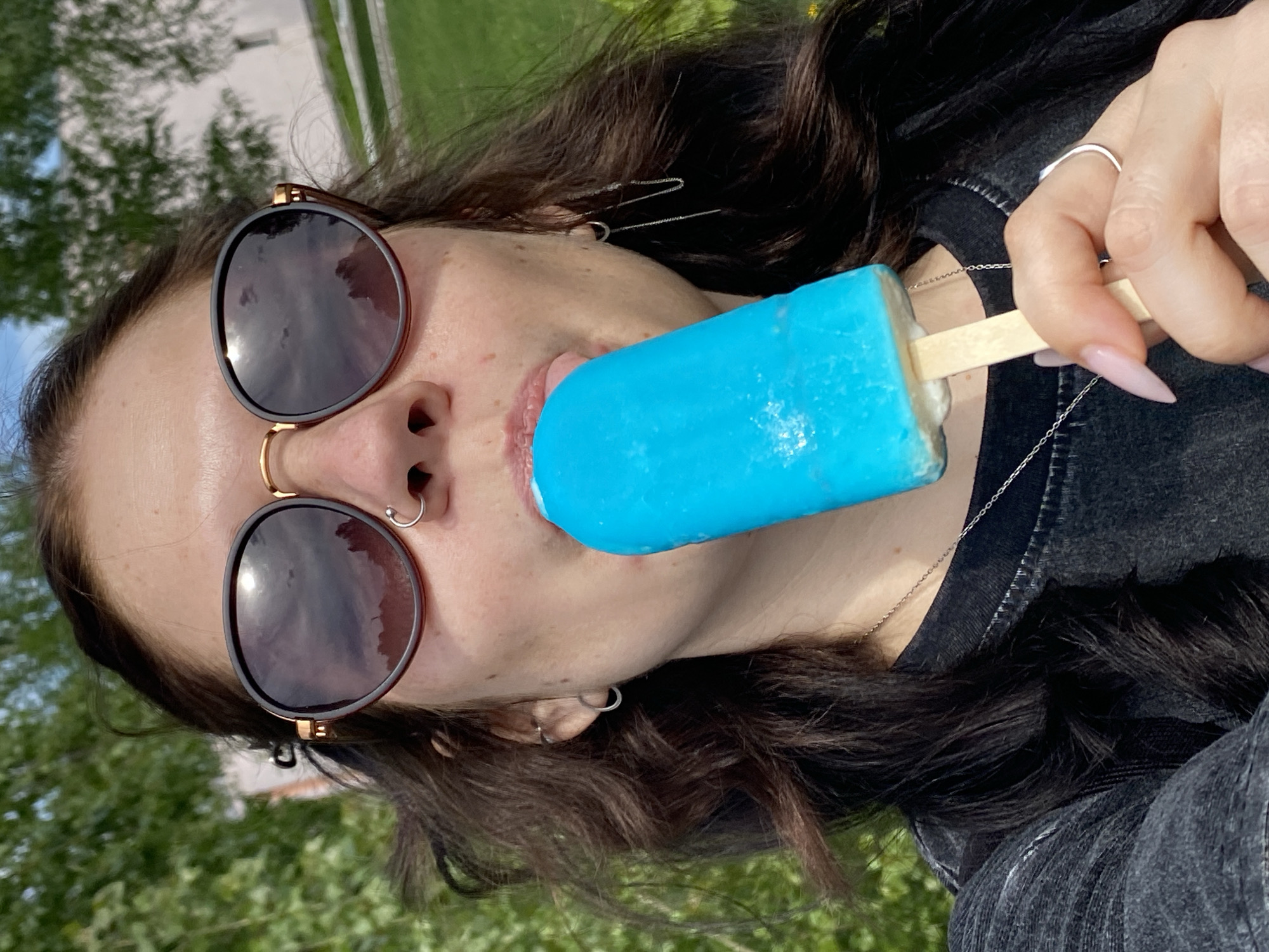 A woman is holding and eating a blue popsicle. She has brown hair, wears sunglasses with round, dark lenses, and has piercings in her nose and ears. She is also wearing a silver necklace and a ring on her finger. The woman is wearing a black shirt, and outside, there is grass and trees visible. The location is Chaikovsky, Russia.