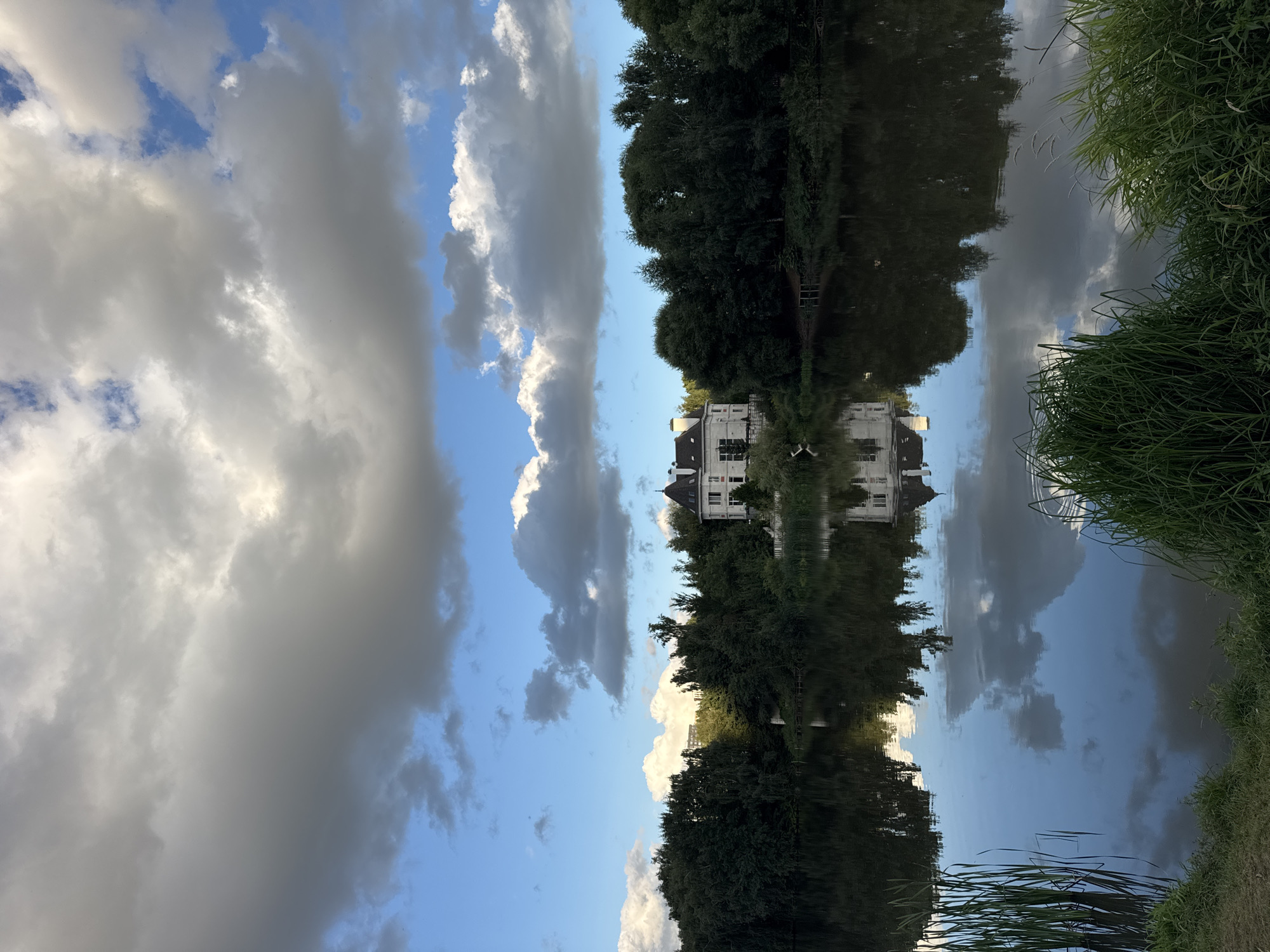 This is an outdoor, eye-level, vertical shot of a tranquil lake in Kommunarka, Russia. A large, white, multi-story house with a dark, conical roof and multiple windows sits nestled amongst dense, dark green trees on the far bank. The house is centrally located on the horizon line. The lake in the foreground reflects the house and the surrounding trees, as well as the dramatic, partly cloudy sky above. The sky is a mix of deep blue and large, billowing white and grey clouds. Reeds and tall green grass line the near bank of the lake. The overall mood is peaceful and serene.