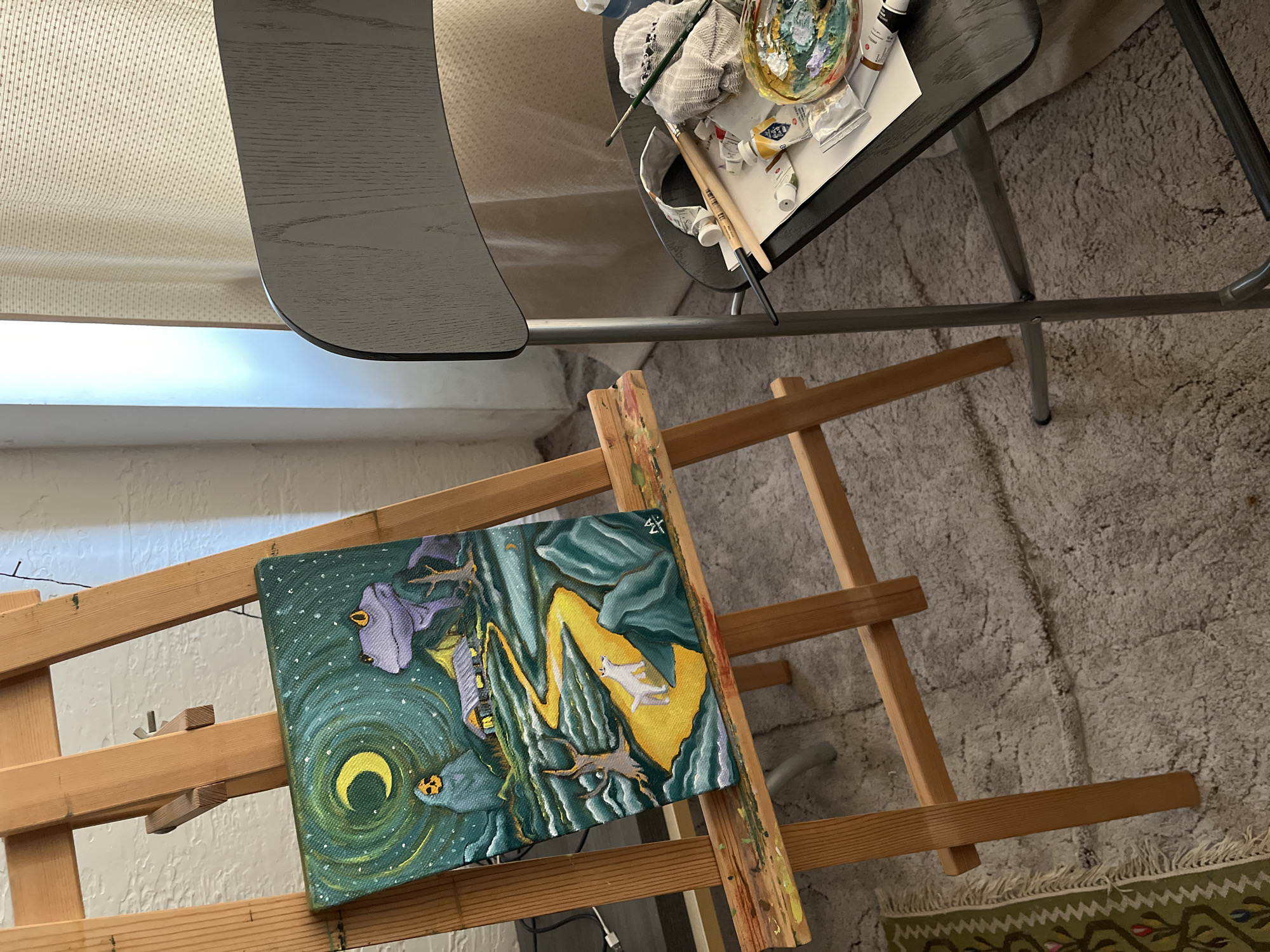 An indoor art workspace is depicted, dominated by a wooden easel holding a vibrant, fantastical painting. The painting, oriented horizontally, showcases a mystical landscape with a dominant green and yellow palette. In the bottom left, a bright crescent moon is encircled by swirling green patterns and dotted with white, suggesting stars. A purple bird-like creature perches on the left, gazing towards the moon, while a white, cat-like or fox-like creature is prominently displayed on a bright yellow, wave-like formation to the right. Below it, a dark, branch-like or antlered figure extends. The background blends swirling greens, blues, and purples, creating an otherworldly atmosphere. A small, stylized signature, resembling "ZS" or "Σ", is visible in the top right corner of the artwork.
To the right of the easel, a dark grey, possibly metal, table is laden with art supplies. These include an artist's palette, various paint tubes (some white, some yellow), several paintbrushes, and a crumpled white rag, indicating active use. The floor is covered with a light-colored, shaggy carpet, and a section of a patterned rug with green borders is visible in the bottom right. The background features an off-white, textured wall, possibly painted brick or stucco. Bright, ambient light streams in from the left, suggesting daytime. A dark, curved furniture piece is partially visible in the upper left, above a light-colored, textured fabric, possibly upholstery or a curtain. A thin, dark cable runs across the carpet near the easel. The overall scene conveys a creative and lived-in artistic environment.