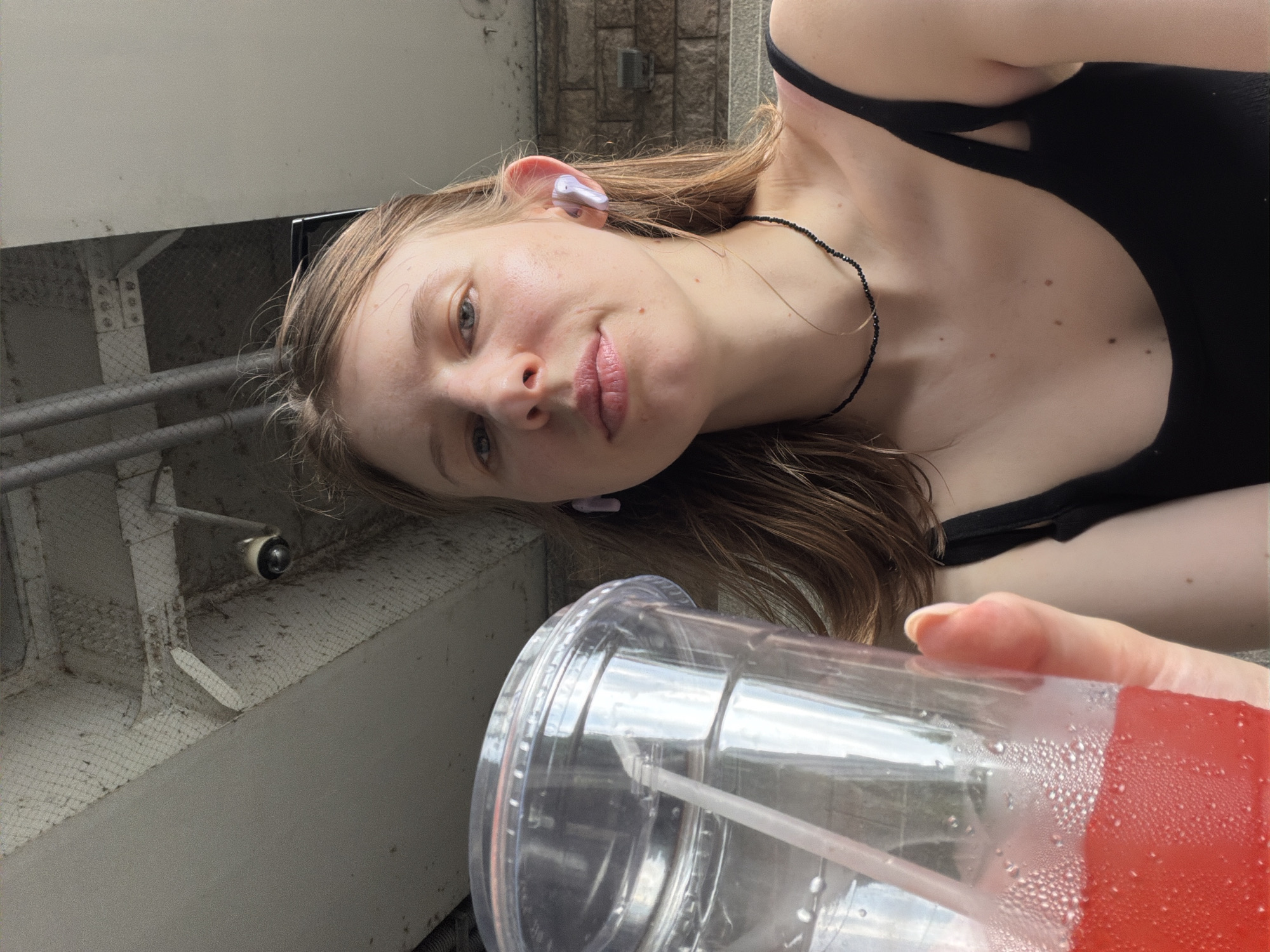 A young woman with light brown hair is holding a clear plastic cup with a red beverage and a straw. She is wearing a black tank top and a beaded necklace. She has purple earbuds in her ears. The background appears to be outdoors, possibly under a bridge or overpass, with a concrete ceiling and some metal pipes visible. There is also a security camera mounted on the ceiling. The overall lighting suggests it is daytime. The location is not specified as Seoul, South Korea.