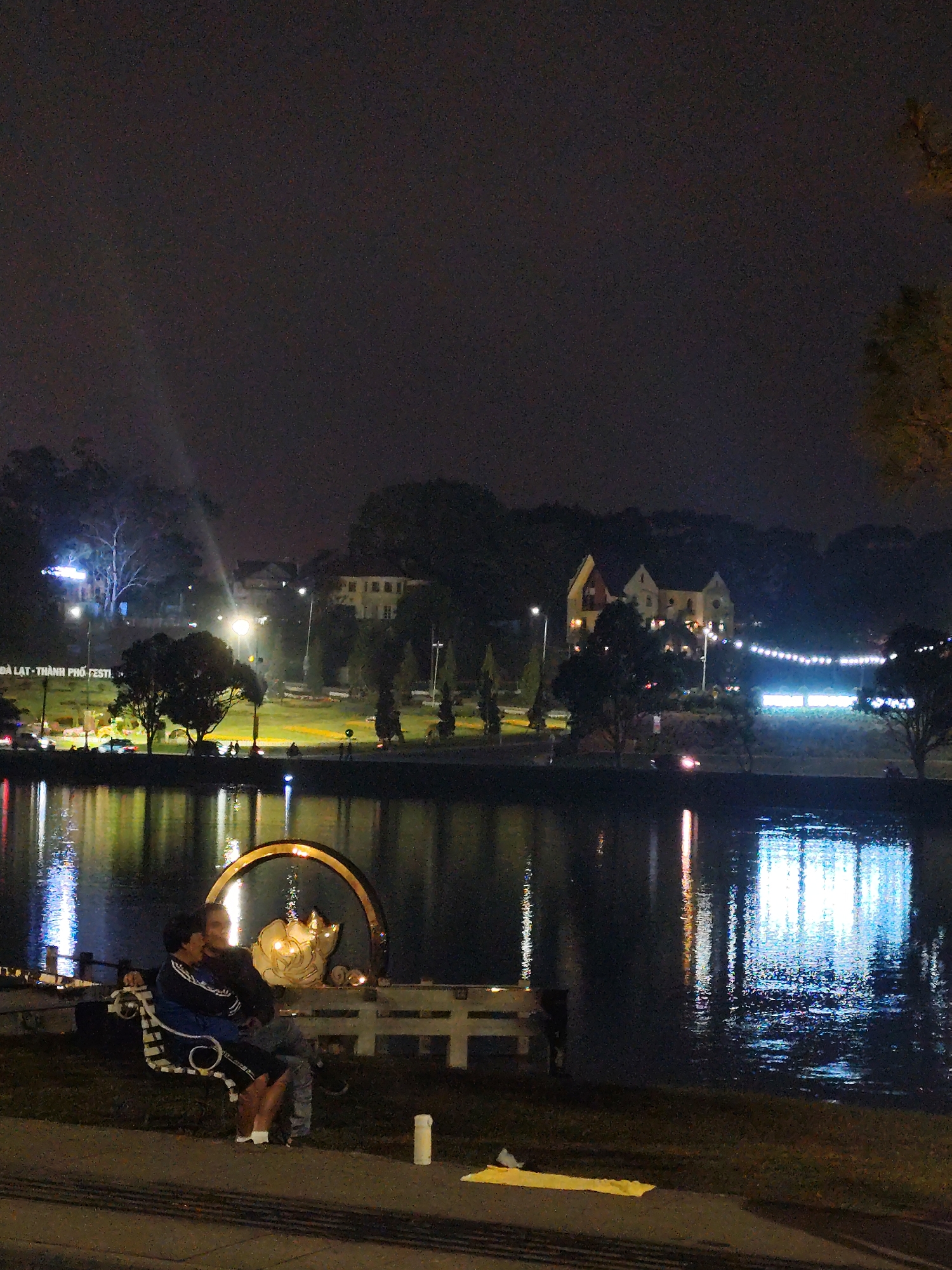 A nighttime view of Xuân Hương Lake in Đà Lạt, Vietnam, features a couple sitting on a white bench in the foreground. Behind them, a decorative circular structure with a lotus flower is illuminated. The lake reflects city lights, buildings, and trees. A white container and a yellow cloth are near the bench. The sky is dark and overcast.