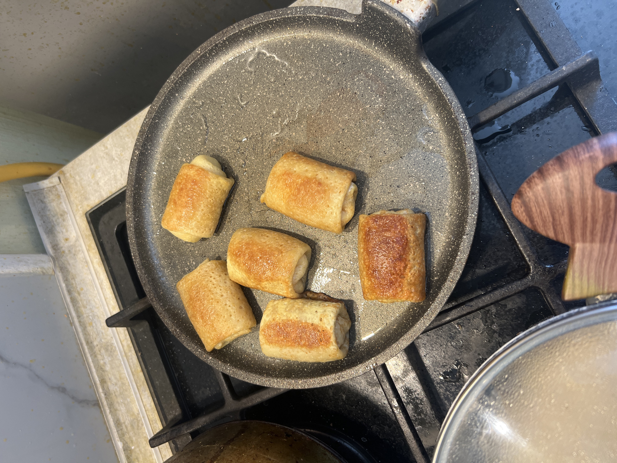 Six golden-brown stuffed crepes are cooking in a circular non-stick frying pan on a black gas stove. The pan has a speckled gray interior and a black exterior. The crepes are cylindrical and appear to be filled with a light-colored substance. The stove has cast-iron grates and a marble-patterned backsplash behind it. To the left, a portion of a large dark pot with a lid is visible. In the foreground, a wooden utensil with a curved handle is partially visible. The scene takes place in a kitchen, likely in Volgograd, Russia.