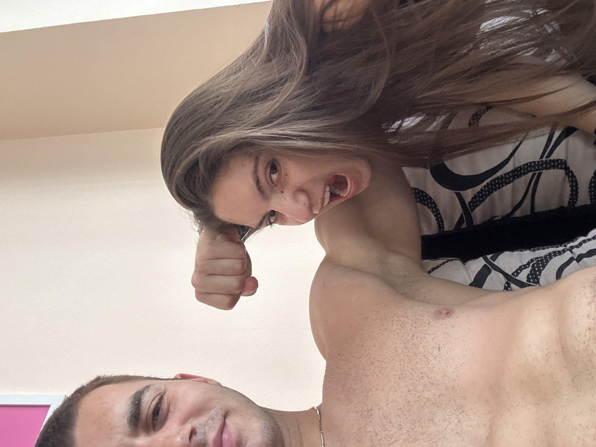 Two individuals, a woman and a man, are captured in a low-angle, close-up selfie, with the camera positioned looking upwards. The woman's face is prominent, her mouth open in a playful or surprised expression, with her long, brown hair cascading around her. She is leaning in, her fist visible near her face. The man's face is at the bottom of the frame, looking upwards and smiling. He is shirtless, and his chest is visible. The background appears to be a bedroom, with a patterned duvet or bedspread visible behind the woman, and a plain, light-colored wall. There are no discernible time of day or weather cues. The overall mood is informal and intimate.
