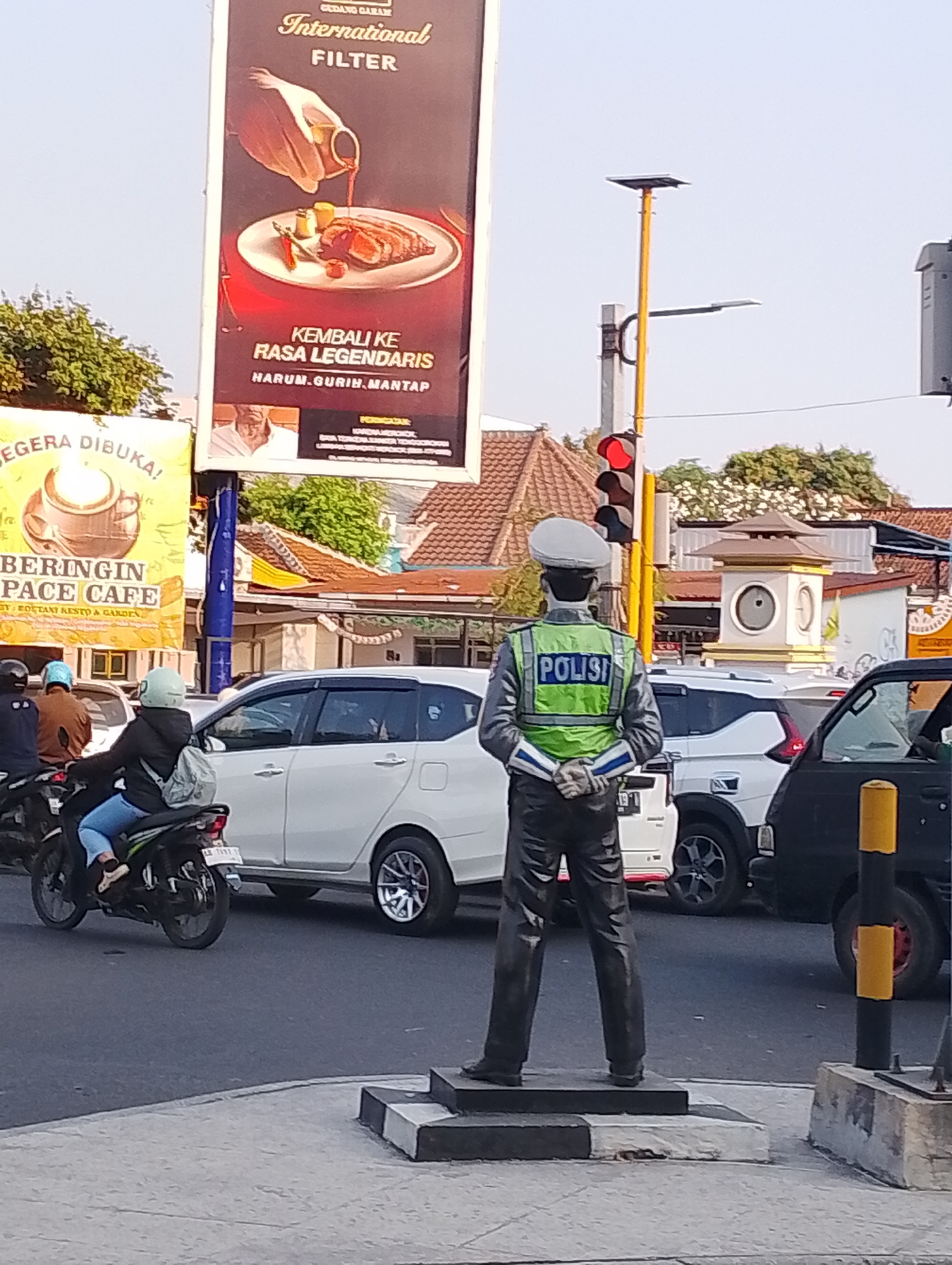 The image shows a statue of a police officer standing at a street intersection in Yogyakarta, Indonesia. The statue is wearing a light blue uniform with a reflective green vest that has "POLISI" printed on it in blue letters. It is standing with its hands clasped behind its back. The statue is positioned on a pedestal in the foreground.
In the background, there is a traffic light, a clock tower, and several buildings with tiled roofs. The street is busy with traffic, including cars and motorcycles. There are also several billboards visible. One billboard advertises "GUDANG GARAM International FILTER" and shows an image of a steak. Another billboard advertises "BERINGIN SPACE CAFE".
The weather appears to be clear and sunny. The overall scene depicts a typical street corner in a bustling Indonesian city.