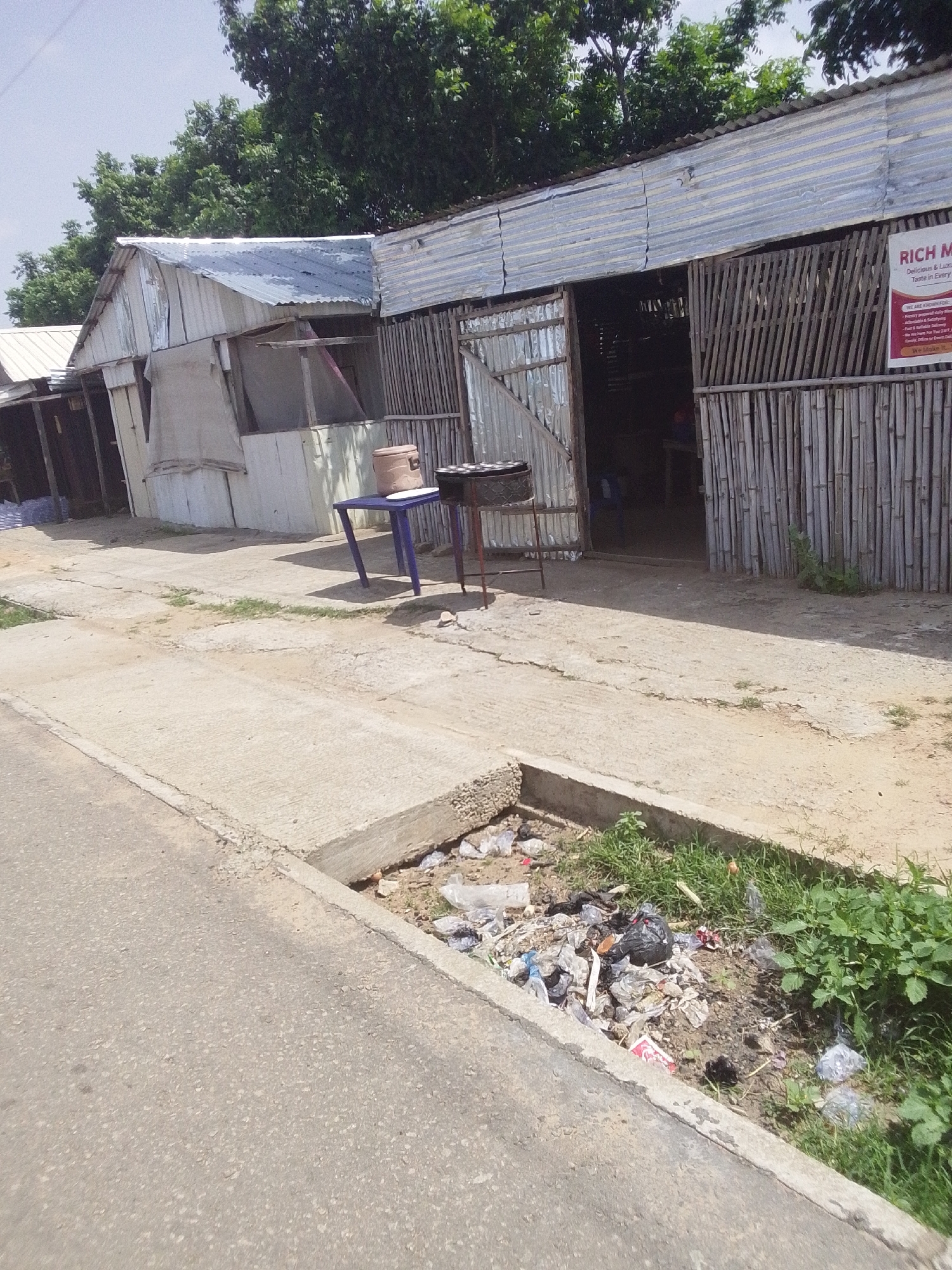 The image shows a roadside scene in Jalingo, Nigeria, featuring a row of informal market stalls or shops. The structures are made of corrugated metal and bamboo, with open fronts suggesting they are places of business. A blue table with a brown pot and a grill is situated outside one of the stalls. A sign with text, including "RICH M," is visible on the facade of one building, indicating a business name and possibly offering food services. The ground in front of the stalls is a mix of concrete and dirt, with a small ditch filled with litter, including plastic bags and other debris, along the edge of the paved road. Lush green foliage is present in the background, suggesting a tropical climate. The overall impression is one of a bustling, informal commercial area.