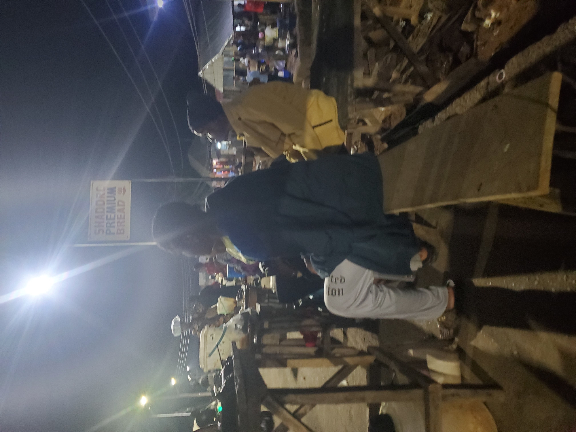 The image depicts a nighttime scene in Gwong, Nigeria, likely a street market or gathering. Several people are present, some of whom appear to be vendors or customers. One person in the foreground is wearing a dark blue jacket and light grey pants with white lettering that reads "Limited Edition." Another individual, further back, is wearing a tan jacket. The scene is illuminated by artificial lights, including a bright light on the left, creating lens flares and casting shadows. In the background, there's a sign that reads "SHADOK PREMIUM BREAD." The setting appears to be outdoors, with what looks like wooden structures and debris suggesting a temporary setup or construction. The overall mood is busy, with a sense of activity despite the darkness. The weather is not discernible from the image.