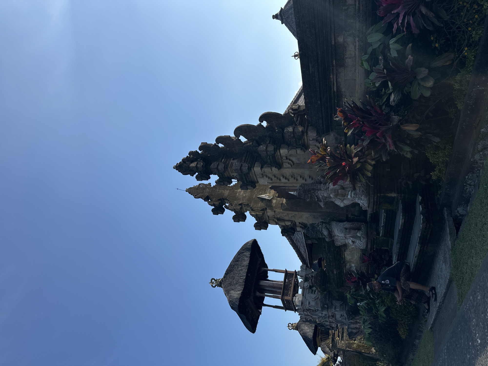 The image captures a traditional Balinese temple entrance, specifically located in Bangli, Indonesia. The architecture is characterized by intricate stone carvings and tiered roofs, common in Balinese religious structures. A prominent feature is a pavilion-like structure with a thatched roof, topped with a decorative crown. In the foreground, a man is seated on stone steps leading up to the temple complex. He is wearing traditional Balinese attire. The surrounding environment includes lush tropical foliage, with plants displaying vibrant red and green hues. The sky is clear and blue, suggesting a sunny day.