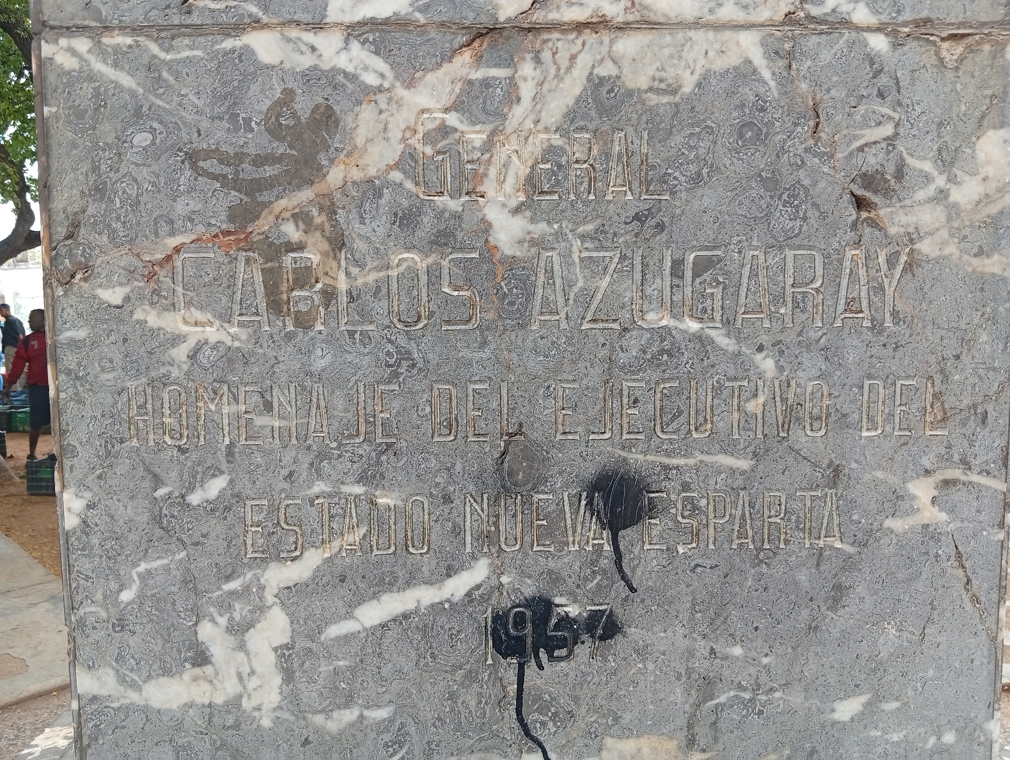 This image shows a marble monument in Juan Griego, Venezuela, bearing an inscription. The inscription reads: "GENERAL CARLOS AZUGARAY HOMENAJE DEL EJECUTIVO DEL ESTADO NUEVA ESPARTA 1957". The marble has natural veins and some areas of discoloration. There is a black spray-painted mark that obscures part of the inscription, particularly the date. In the background, to the left of the monument, a tree with green foliage is visible. Also in the background, two people are partially visible, one wearing a red shirt and another in dark clothing. The ground appears to be paved. The overall scene suggests an outdoor public space.