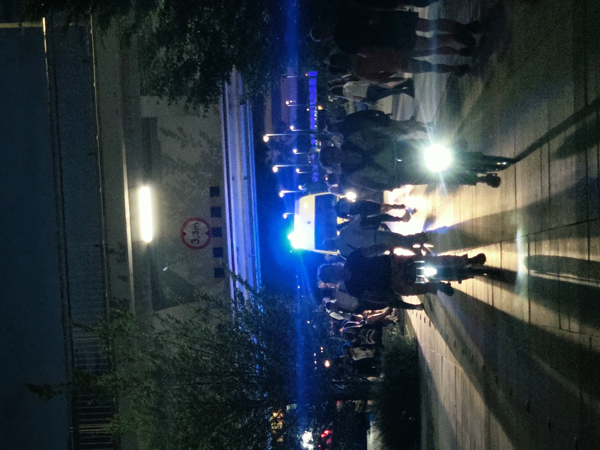 In Warsaw, Poland, at night, a crowd of people is gathered near an overpass. A few individuals are riding bicycles with bright headlights that cast long shadows on the paved walkway. An ambulance with flashing blue lights is visible in the background. Trees line the sides of the walkway, and streetlights illuminate the scene. A sign indicating a height restriction of 3.3 meters is attached to the overpass.