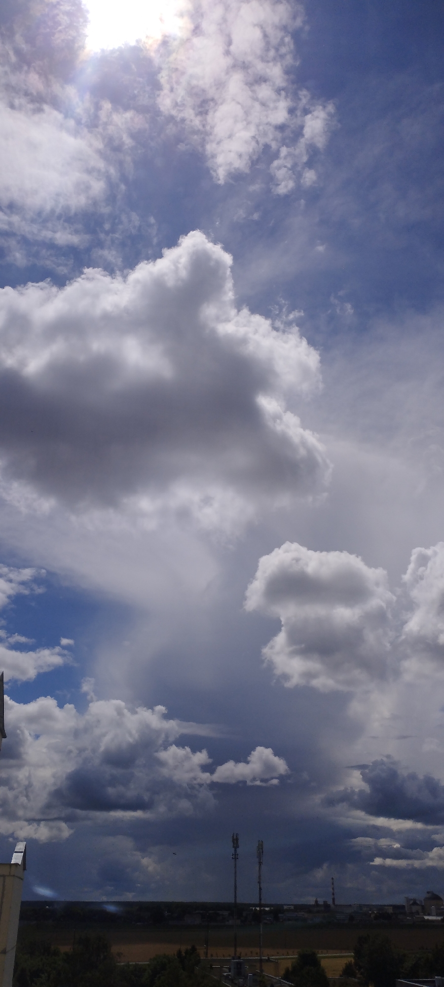 This is a vertical, outdoor, daytime photograph taken from an elevated vantage point, likely a rooftop, overlooking a landscape with a dramatic sky. The scene is dominated by a vast expanse of sky filled with various types of clouds. The upper portion of the image features bright, white cumulus clouds, with some appearing dense and textured, against a bright blue sky. A strong light source, possibly the sun, is visible in the upper left quadrant, causing lens flare and washing out some of the cloud detail.
As the eye moves down, the clouds become larger and darker, transitioning into grey and deep grey formations, suggesting the presence of storm clouds or heavy overcast. These darker clouds create a stark contrast with the lighter sky above.
The lower portion of the image shows a distant horizon line. Silhouetted against the darker sky is a town or city, with various structures visible. These include what appear to be communication towers with antennas, a factory or industrial complex with chimneys, and several buildings. A thin, horizontal line of trees separates the town from the sky. In the very foreground, the edge of a light-colored building or structure, possibly a rooftop with a parapet, is visible on the left side of the frame. The overall impression is of a dynamic sky over a semi-urban or industrial landscape in Skidzieĺ, Belarus. No people are visible in the frame.