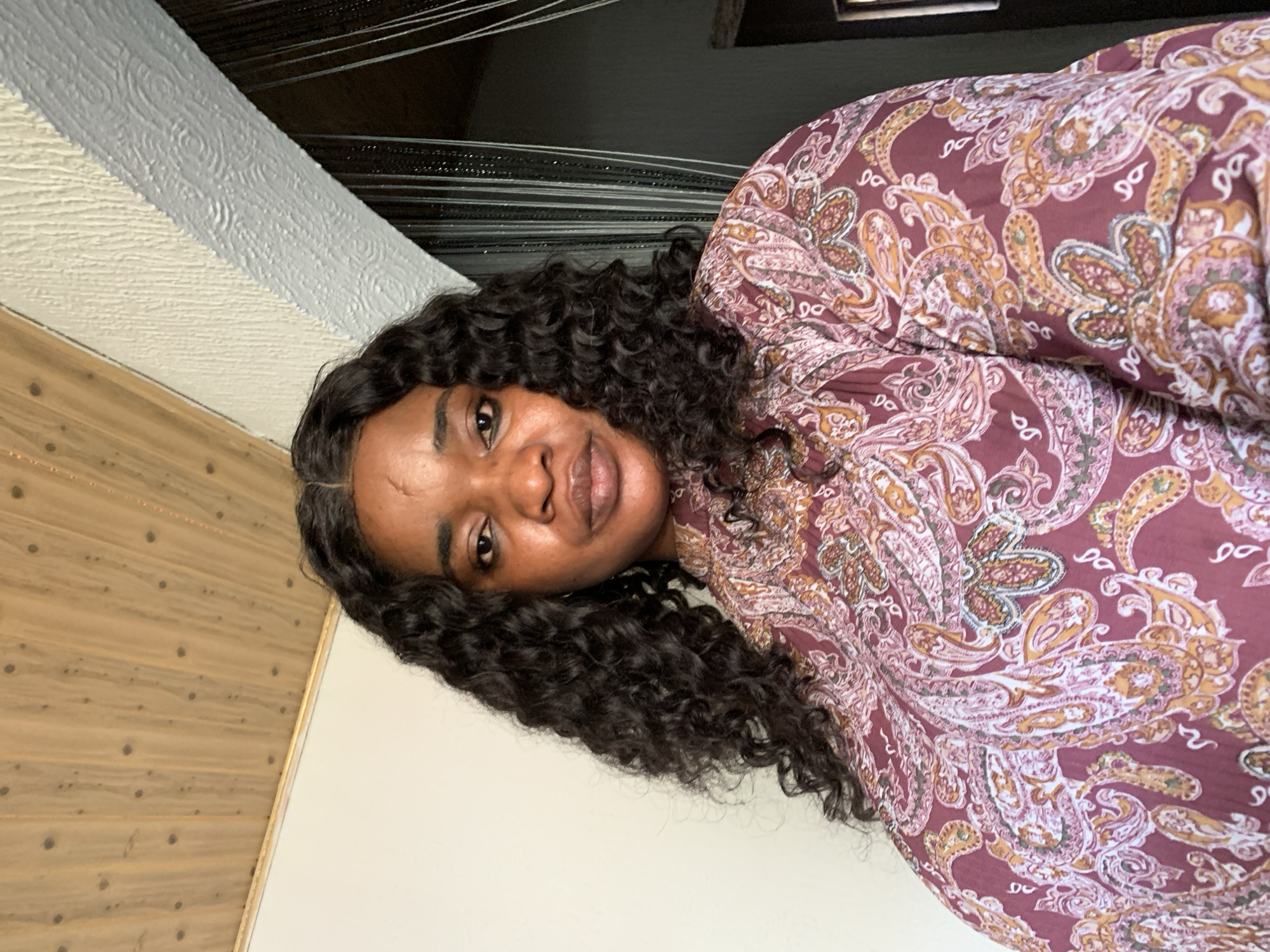 A medium shot shows a woman facing forward, with her head and shoulders filling most of the frame. She has dark, curly hair that falls around her shoulders. Her skin appears to have some texture and slight blemishes. She is wearing a burgundy-colored shirt with a paisley pattern in shades of pink, white, and orange. The background is divided into different textures: on the left, a white wall; above, a wooden ceiling with visible planks and knots; and on the right, a textured wall in a light color, with vertical dark curtains or blinds hanging in the background. The lighting suggests an indoor setting. This image was taken in Barakin Akawo, Nigeria.