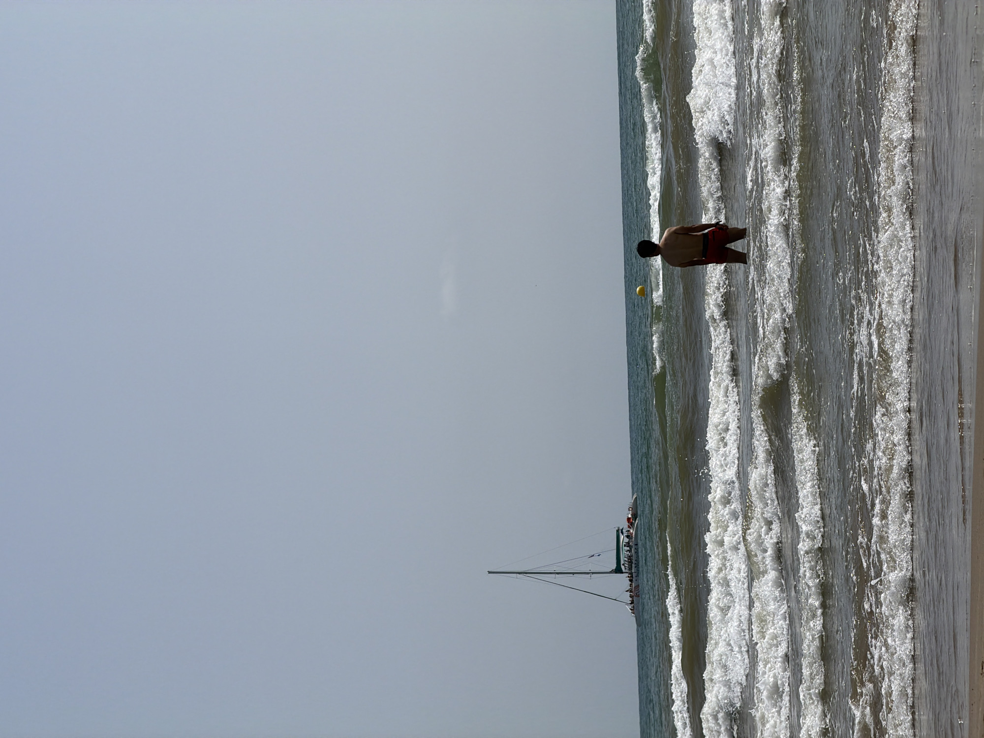 In an outdoor, daytime scene at a beach, a man stands in the ocean. He is facing away from the camera, wearing red shorts. The water is choppy with white-capped waves. In the distance, a white catamaran with people on board sails across the blue-green ocean under a hazy sky. A yellow buoy is visible in the water to the left of the man. The sand of the beach is visible in the foreground.