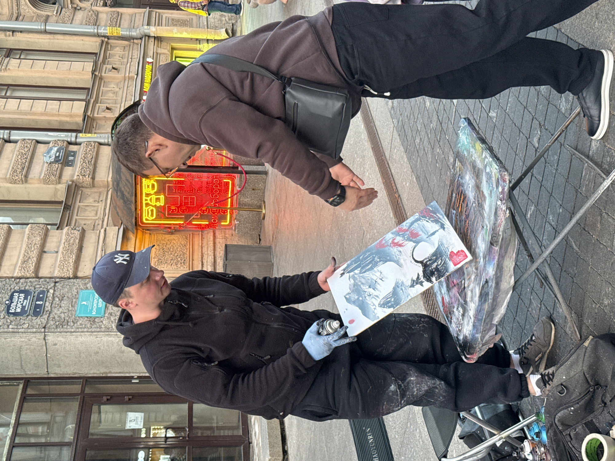Two men are in an outdoor urban environment, possibly Saint Petersburg, Russia, engaged in street art. One man, wearing a black hoodie and a New York Yankees baseball cap, holds a spray can in his right hand, which is covered by a light blue glove, and is applying spray paint to a painting of a landscape with mountains and birds. The painting is propped up on a folding table, which is covered with plastic sheeting and appears to be splattered with paint. The man is wearing black pants that are also splattered with paint. He is looking down at the painting. The second man, wearing a brown hoodie and glasses, is standing to the right of the table, also looking at the painting and appears to be interacting with the first man. He has a black messenger bag slung over his right shoulder and is wearing black pants and black sneakers. In the background, a building with a historical facade and arched windows is visible. There are also signs on the building, one of which reads "БОЛЬШАЯ МОРСКАЯ" which translates to "Bolshaya Morskaya Street" a prominent street in Saint Petersburg. Another sign in teal reads "ВОЛОНТЕРСКИЙ ЦЕНТР" indicating a volunteer center. A neon sign with red illumination is visible on the building. In the foreground to the left, there is a folding chair and several spray cans and bags. The ground is paved with cobblestones.