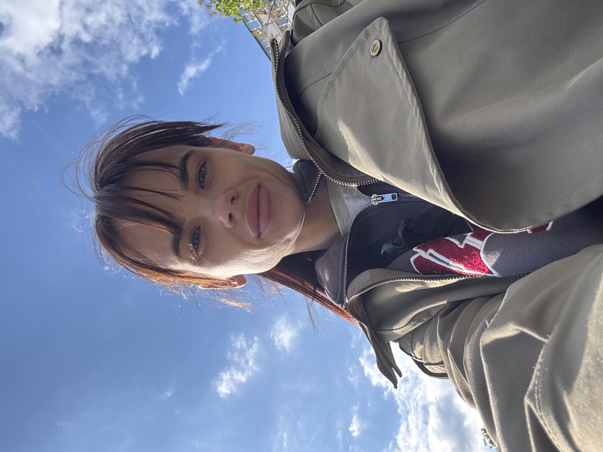 A low-angle, close-up selfie photograph shows a woman with short brown hair and fair skin. She is wearing a light khaki-colored jacket with a zipper. Under the jacket, a gray sweatshirt with a large, red "W" design is visible. The background is a bright blue sky with scattered white clouds.
No location or city-specific details are discernible from the image.
