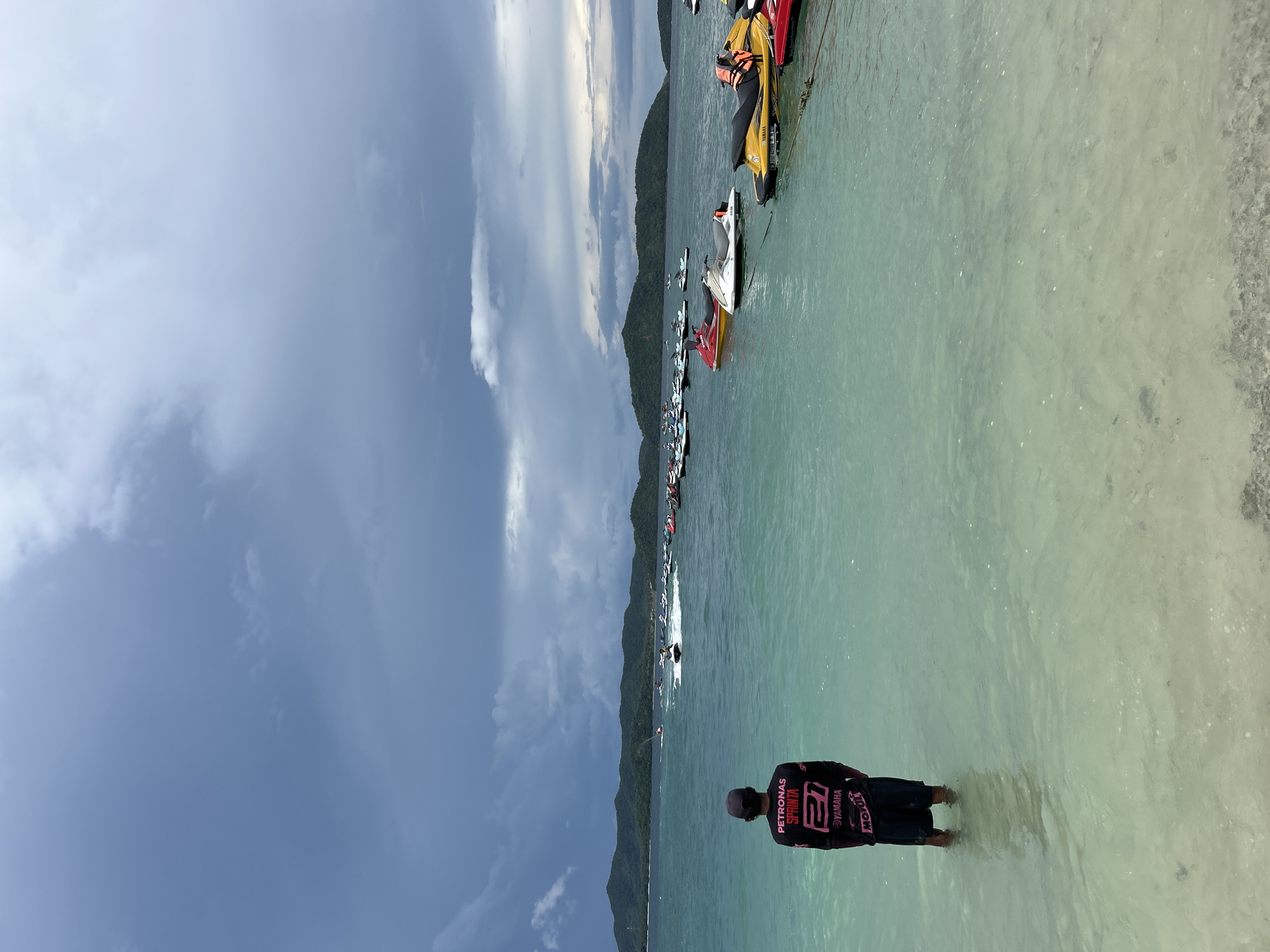 A person stands in shallow, clear turquoise water facing away from the camera, looking out at a body of water dotted with numerous jet skis and distant, green, mountainous islands under a cloudy sky. The person is wearing a dark long-sleeved shirt with pink and white writing, including "PETRONAS SPRINTS" and "YAMAHA," and dark shorts. Several jet skis are moored in the water closer to the shore, some in bright colors like yellow, red, and white, each with an orange life vest. Further out, more jet skis are visible, some in motion, creating white wakes on the water. The background features a range of green hills or mountains with a cloudy, dramatic sky overhead. The scene appears to be a coastal area in Phru Nai, Thailand, likely a popular spot for water sports.