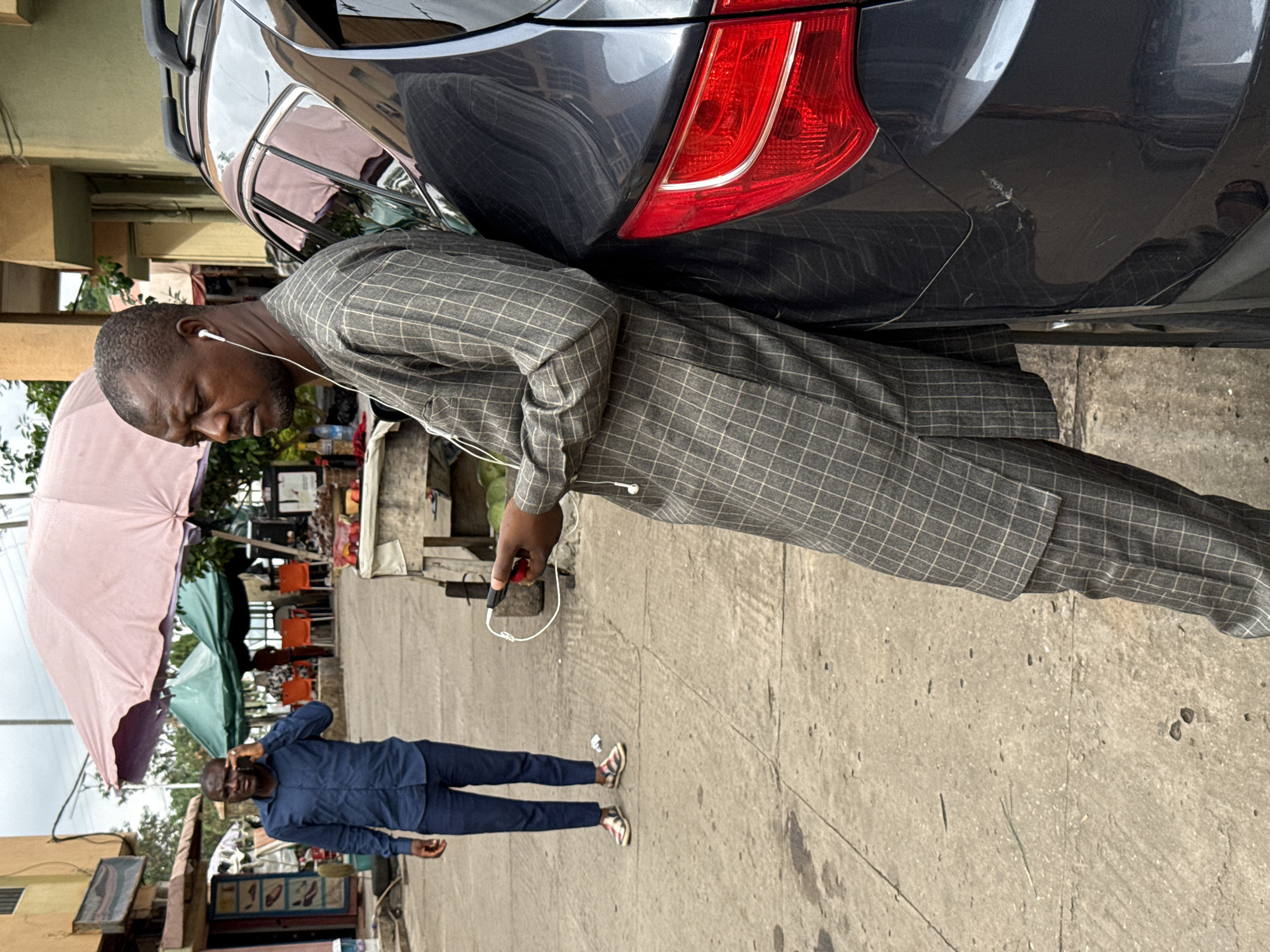 The image depicts a street scene in Abuja, Nigeria. A man in a gray checkered suit is leaning against the rear of a dark gray car, seemingly resting or waiting. He is wearing earphones and holding a device in his hand, possibly a phone or remote. The car has a visible red taillight and minor scratches on the bumper. In the background, another man in a blue shirt and dark trousers is seen talking on his phone while standing on the sidewalk. There are street vendors with stalls covered by umbrellas, suggesting a commercial area. The surroundings include buildings with visible signage and street furniture, indicating a moderately busy urban environment. The ground is concrete and shows signs of wear and staining.