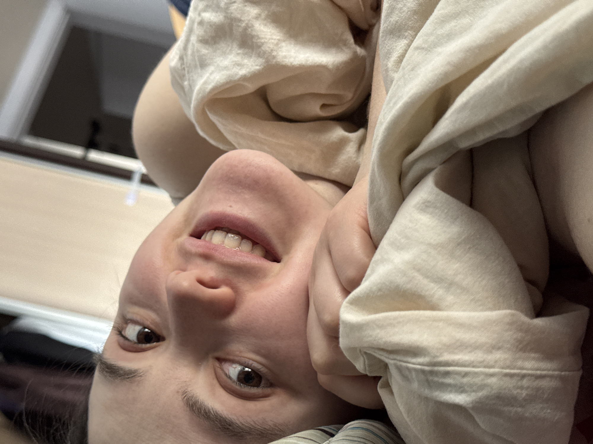 The image shows a person lying down, with their head upside down in the frame. The person is holding a light-colored garment. The environment includes a window and possibly bedding. Location information is not discernible.