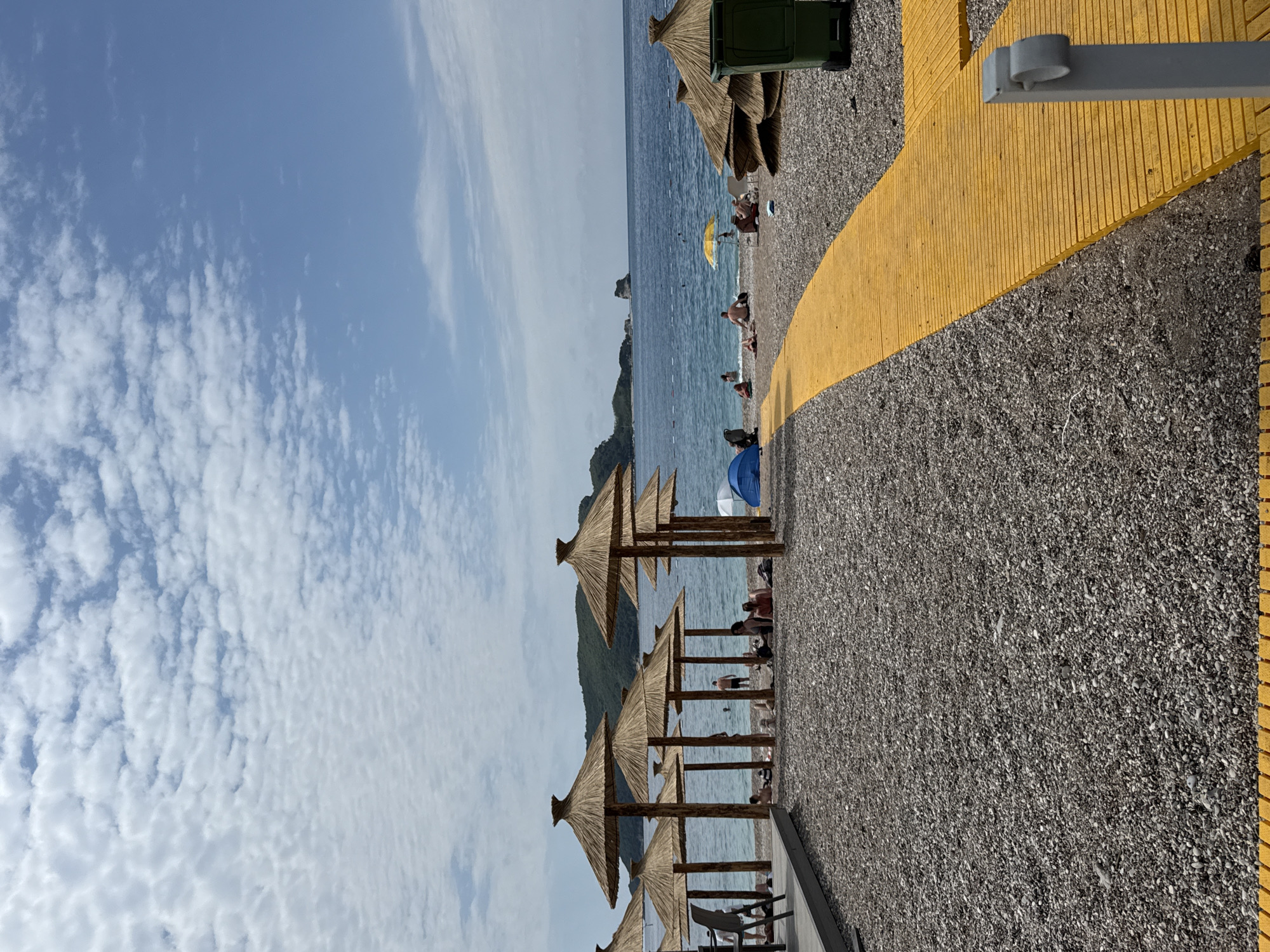This image captures a vibrant and sunny beach scene, likely taken during the day. The setting is a pebbly beach bordering a calm, blue sea, with distant land features, possibly hills or islands, visible on the horizon.
The sky, occupying a significant portion of the upper frame, is a clear blue with scattered white, fluffy clouds, indicative of good weather.
In the foreground and extending along the beach, numerous traditional straw-thatched beach umbrellas (palapas) are neatly lined up, casting short shadows beneath them. Several individuals are visible on the beach, some relaxing under these umbrellas on chairs, while others are closer to the water's edge or in the sea, presumably swimming or wading. A bright yellow beach umbrella and a blue beach tent or umbrella are also noticeable among the crowd.
A distinctive bright yellow pathway, appearing to be textured or ribbed, curves along the right side of the beach, providing a clear walking path. In the immediate foreground, a grey, L-shaped fixture is attached to this pathway, possibly part of a railing or a light post base. A dark green trash receptacle is partially visible near the upper right, tucked under one of the straw umbrellas.
The sea appears calm with faint lines, possibly indicating swimming lanes or areas. The overall atmosphere is one of leisure and relaxation, typical of a summer day at the beach. There is no visible text in the image.