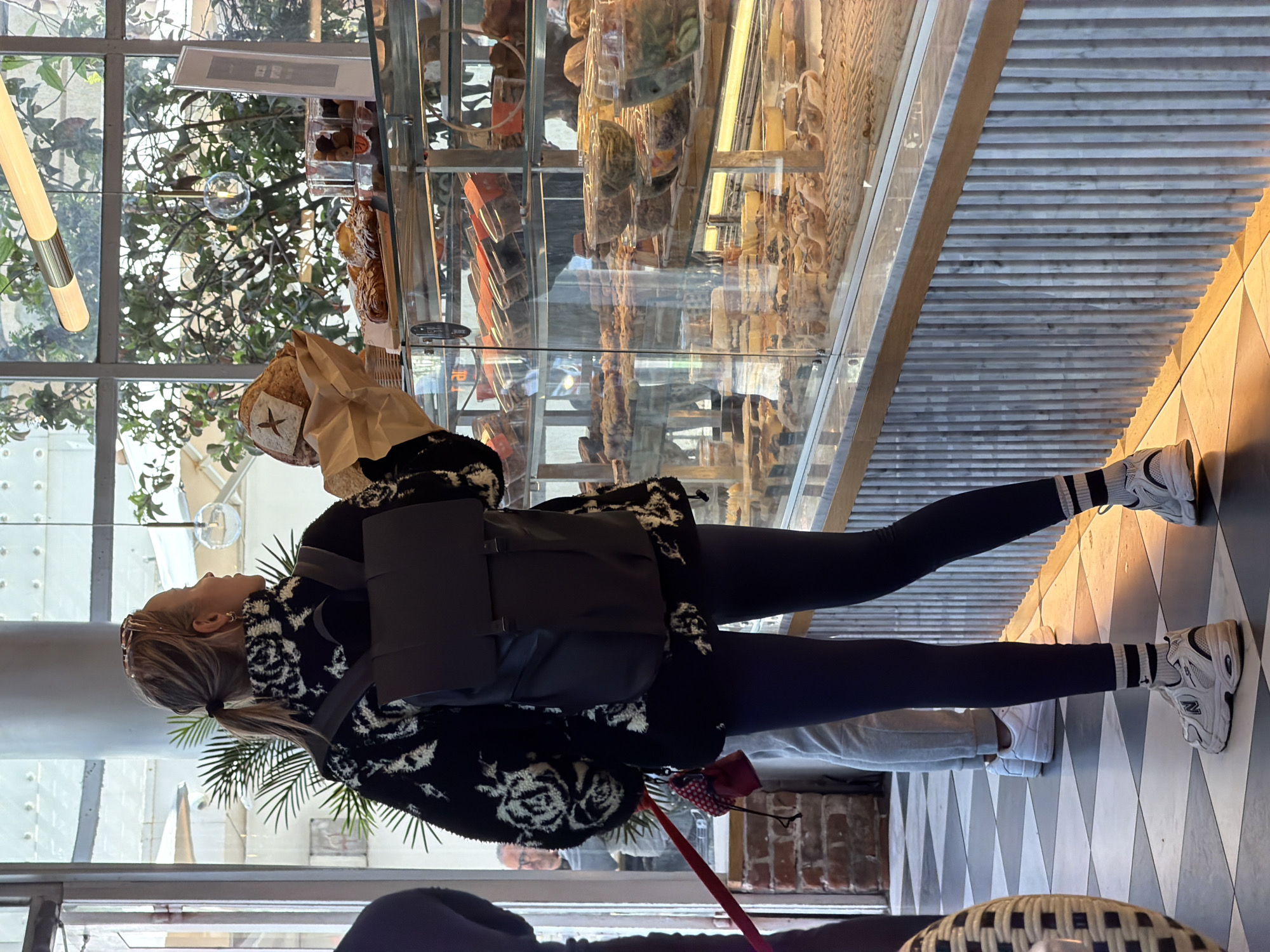 The image shows a woman standing in what appears to be a bakery in Tel Aviv, Israel. She is holding a paper bag containing bread and is wearing a black and white patterned jacket, black leggings, and white sneakers with black stripes on the socks. She has a backpack on and her hair is tied back in a ponytail. A dog leash is visible near her, suggesting she may be with a dog. In the background, there is a display case filled with various baked goods. Through the large window, trees and other elements of the urban environment are visible, including light fixtures and architectural details. The interior of the bakery features a checkered floor. Other individuals are present in the background, contributing to the overall ambiance of the location.