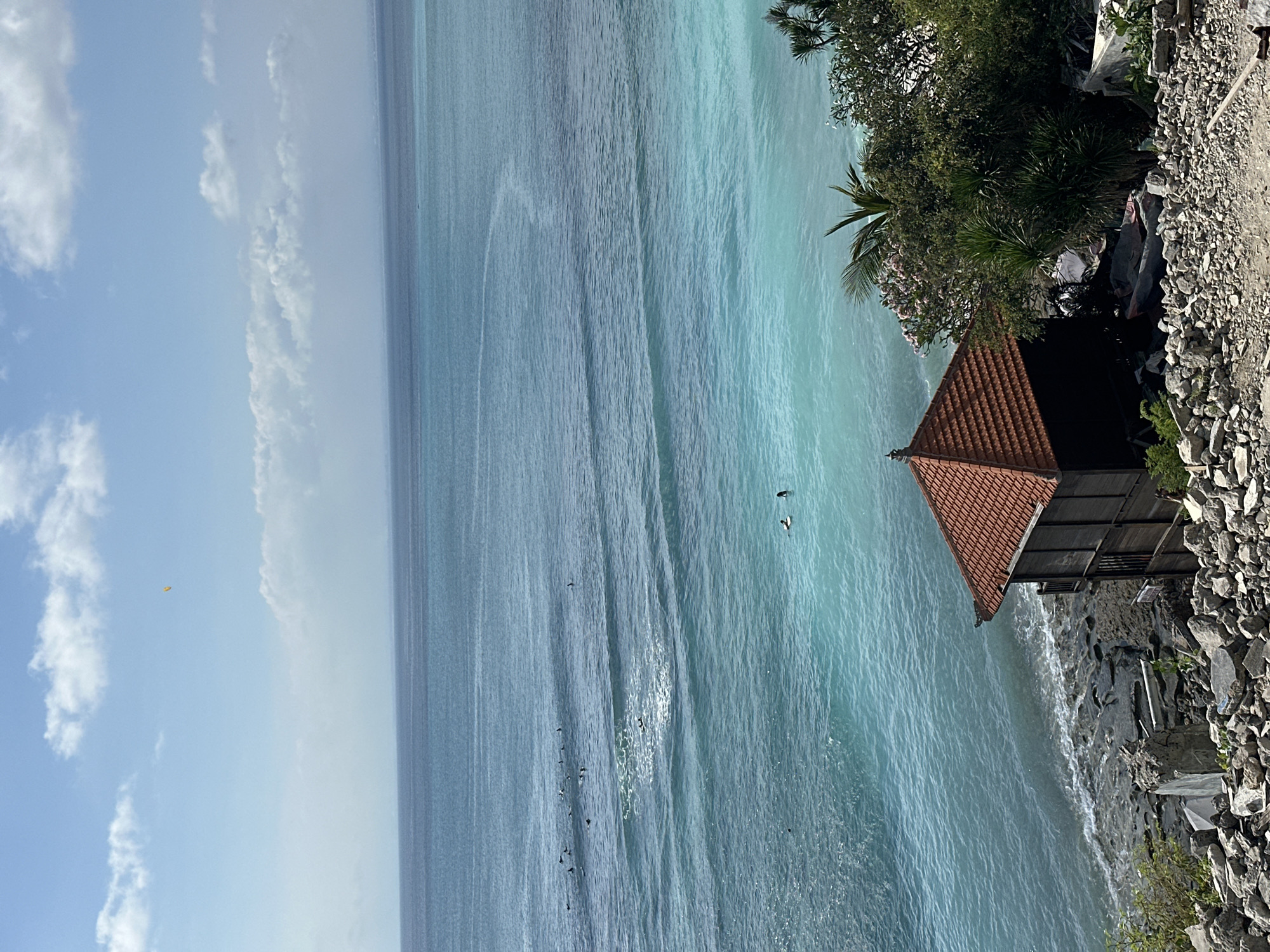 An eye-level, outdoor, high-angle, long shot shows the ocean with several surfers and two individuals on surfboards in Kuta Selatan, Indonesia. The water is a clear turquoise color, with a few ripples and waves. In the foreground, there is a small, dark wooden structure with an orange tile roof, surrounded by rocks and debris. Lush green trees and bushes with pink flowers are visible behind the structure. The sky is blue with scattered white clouds, and a tiny yellow kite is visible in the distance. The overall scene is peaceful and tropical, with a mix of natural beauty and man-made structures.