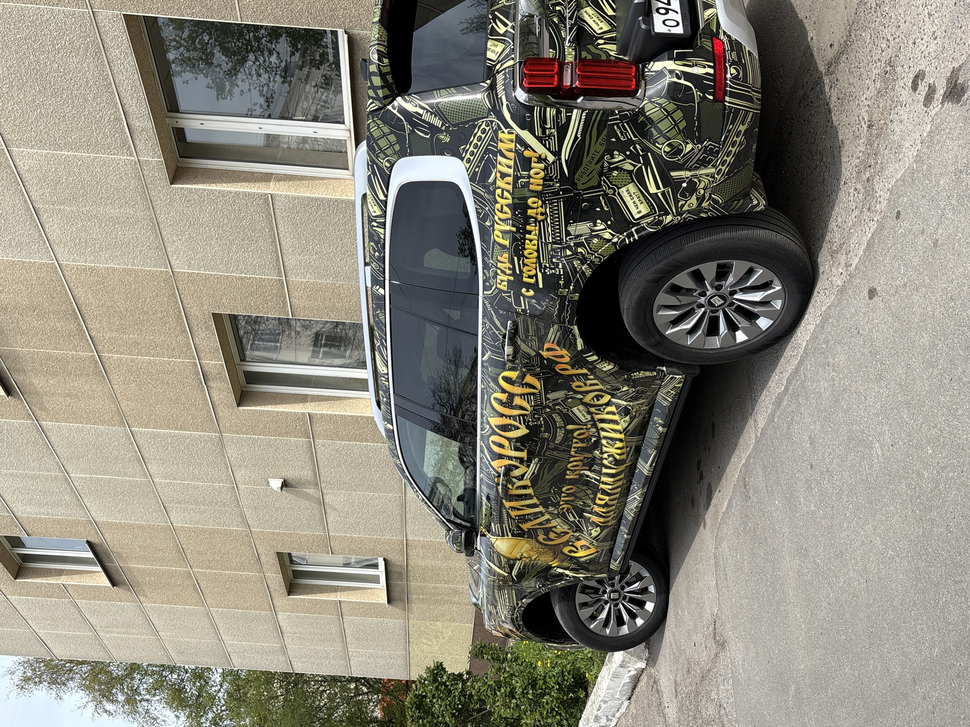 The image shows a parked vehicle with a distinctive military-themed camouflage wrap, partially obscuring an adjacent building, likely taken in Veliky Novgorod, Russia based on the license plate. The wrap includes illustrations of weapons and text written in Russian, including phrases like "Be Russian from head to toe" and "What is the strength, brother?". The vehicle is parked on an asphalt surface alongside a building with multiple windows, some of which reflect the sky. A few trees and shrubbery are visible near the bottom of the image. The license plate is visible, but the numbers are only partially readable.