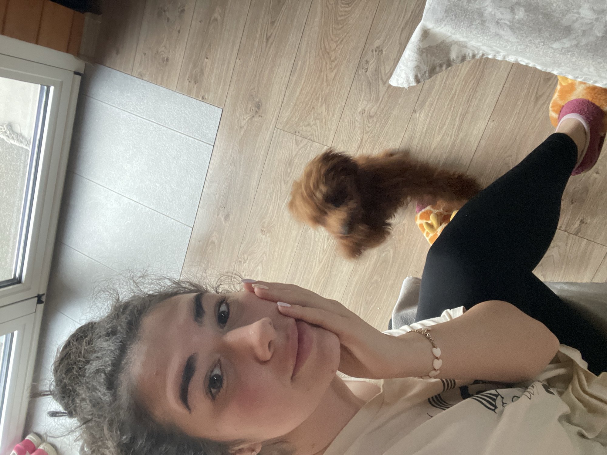 A woman is taking a selfie indoors, in what appears to be an apartment or house. The location cannot be confirmed to be Sofia, Bulgaria based on the visual information provided. The woman has curly hair and is wearing a light-colored top. She has her hand near her face. A small brown dog is on the floor near her. She is wearing black leggings and pink slippers with an orange pattern. The floor is a mix of light brown wood and grey tile. A window is visible on the left side of the frame.