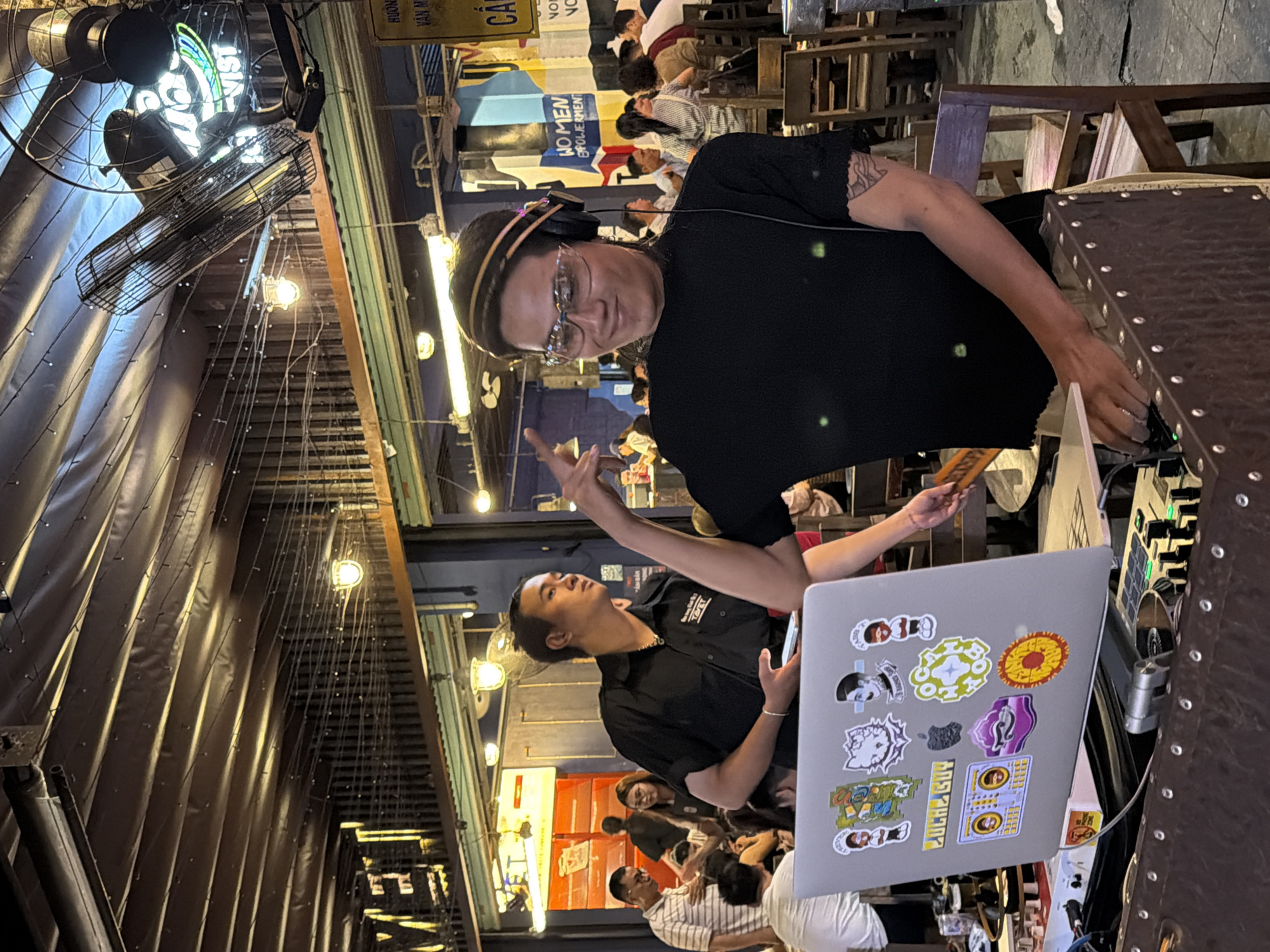 The image captures a vibrant scene at what appears to be an outdoor or semi-open-air establishment in Thuận Hóa Ward, Vietnam. The primary subject is a person wearing headphones and clear-lensed glasses, facing the camera and smiling. They are dressed in a black t-shirt and appear to be a DJ, with a laptop adorned with various stickers in front of them, placed on a DJ console. The laptop displays a collection of colorful stickers, including cartoon characters and a black Apple logo.
Behind the DJ, another individual in a black collared shirt is visible, holding a smartphone. The background is populated with numerous people seated at tables, suggesting a lively social gathering or dining environment. The setting is dimly lit, with overhead string lights and industrial ceiling fans adding to the ambiance. Wooden chairs and tables are prominent elements of the environment, along with metal fixtures and exposed structural elements. The overall impression is that of a casual, energetic evening at a popular spot in Thuận Hóa Ward.