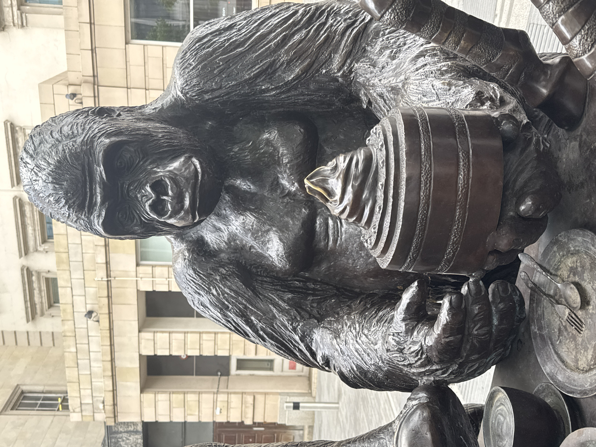 A bronze sculpture of a gorilla is located outdoors, with a backdrop of a multi-story building featuring light-colored stone cladding. The sculpture shows the gorilla holding a tiered object, possibly a cake, with what appears to be frosting on top. Tableware, including a plate, utensils (fork and spoon), and cups, are placed around the gorilla's hands. The location is an urban setting within a city of Unknown, Unknown.