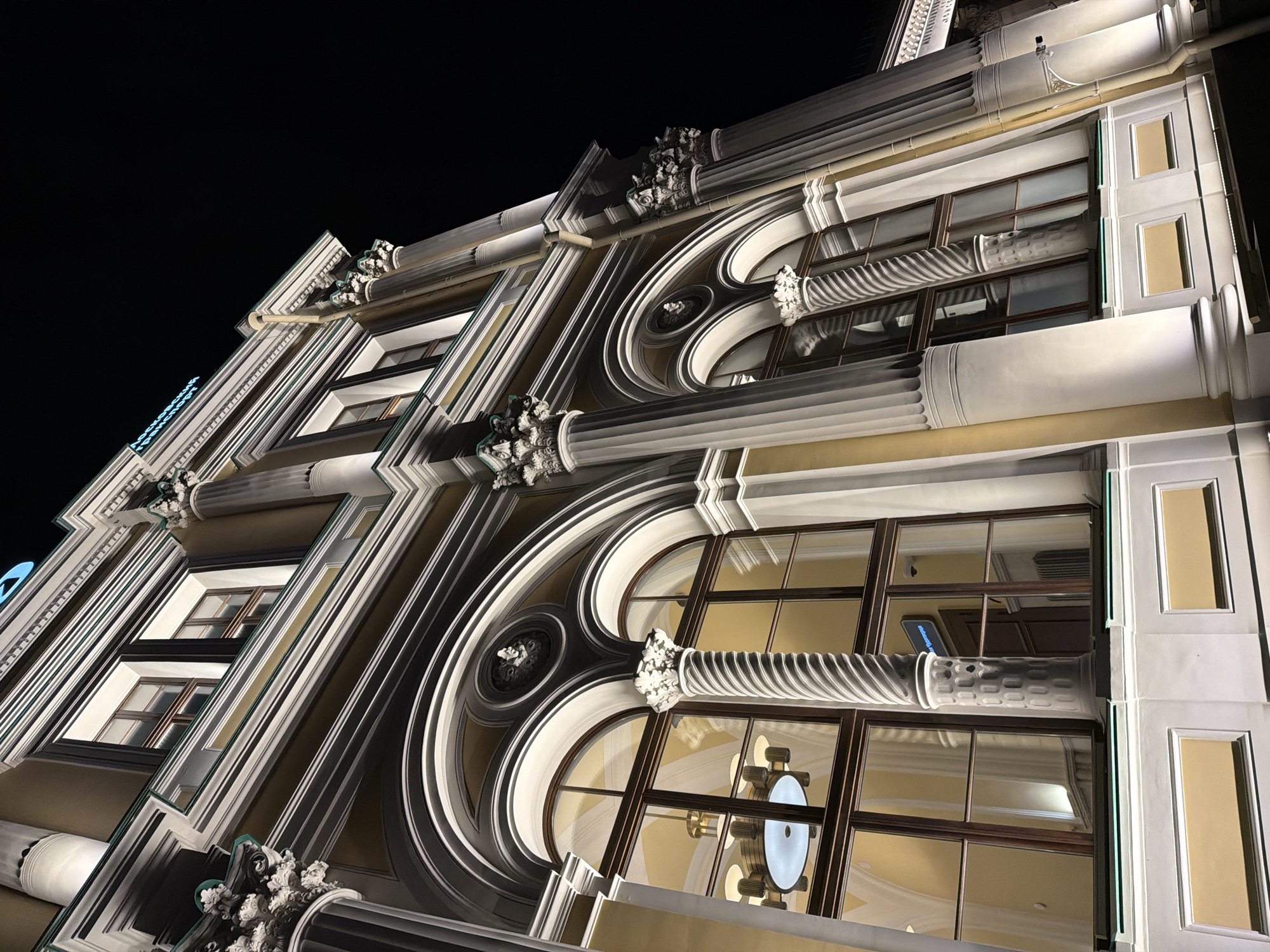 This is a low-angle, night-time, full-frame shot of the Moscow City Duma building in Moscow, Russia. The facade of the building is illuminated by artificial lights, highlighting its neoclassical architecture. Large, arched windows with dark wooden frames are visible, with a chandelier reflecting in one of them. The building features prominent columns with ornate capitals and decorative elements. A neon sign with Cyrillic lettering is visible at the top of the building. The overall scene is one of grandeur and historical significance, set against the dark night sky.