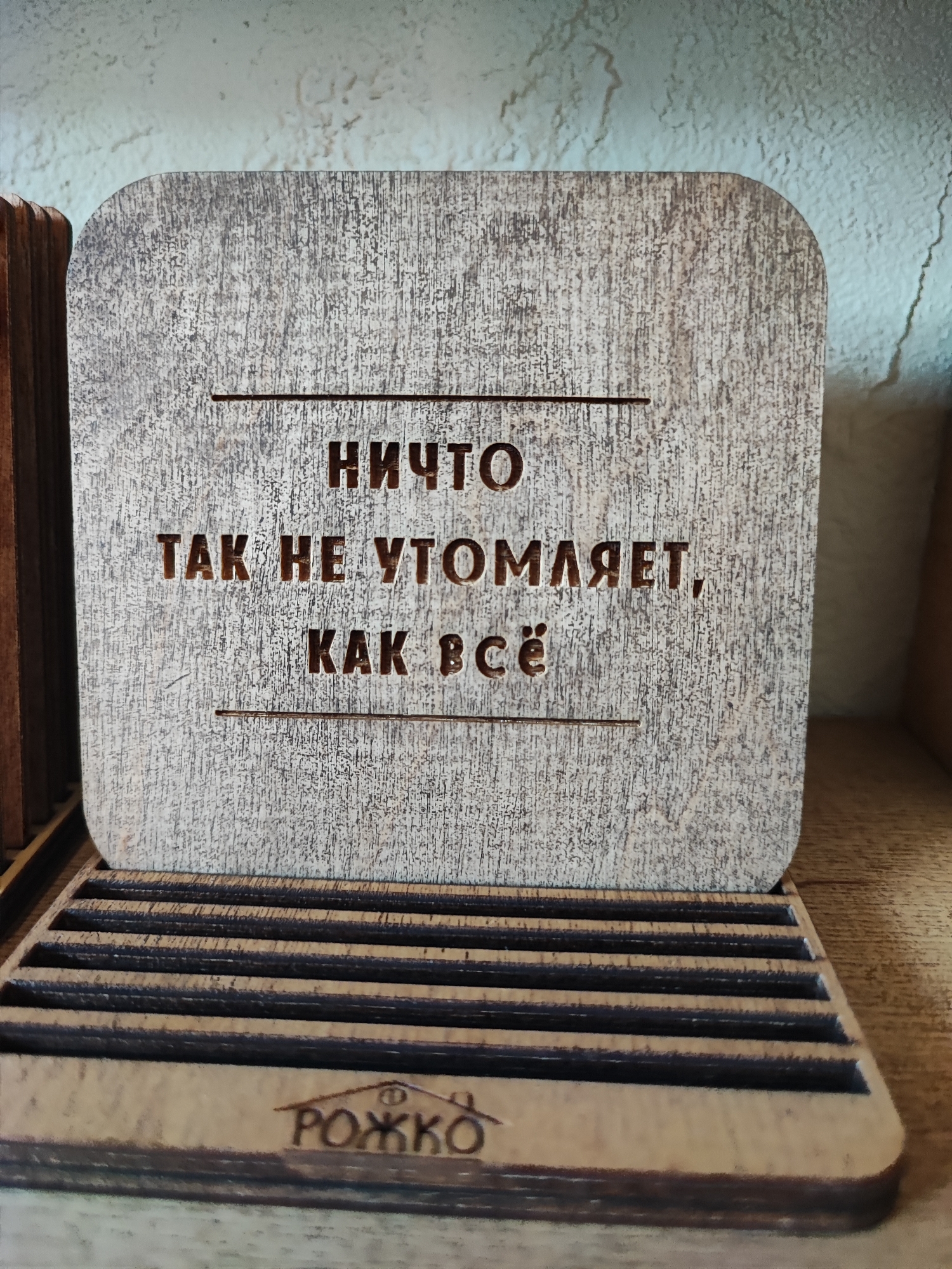 The image shows a wooden quote block with Russian text and a wooden organizer. The quote block is square with rounded corners and is placed in a wooden organizer with multiple slots. The text on the quote block reads "НИЧТО ТАК НЕ УТОМЛЯЕТ, КАК ВСЁ" which translates to "Nothing is as tiring as everything." Below the quote block, on the organizer, is a logo that appears to be a house with the letters "РОЖКО" underneath. The background is a textured wall. The scene is likely indoors, possibly in a home or shop, and no specific location in Pyatigorsk, Russia can be determined from the image alone.