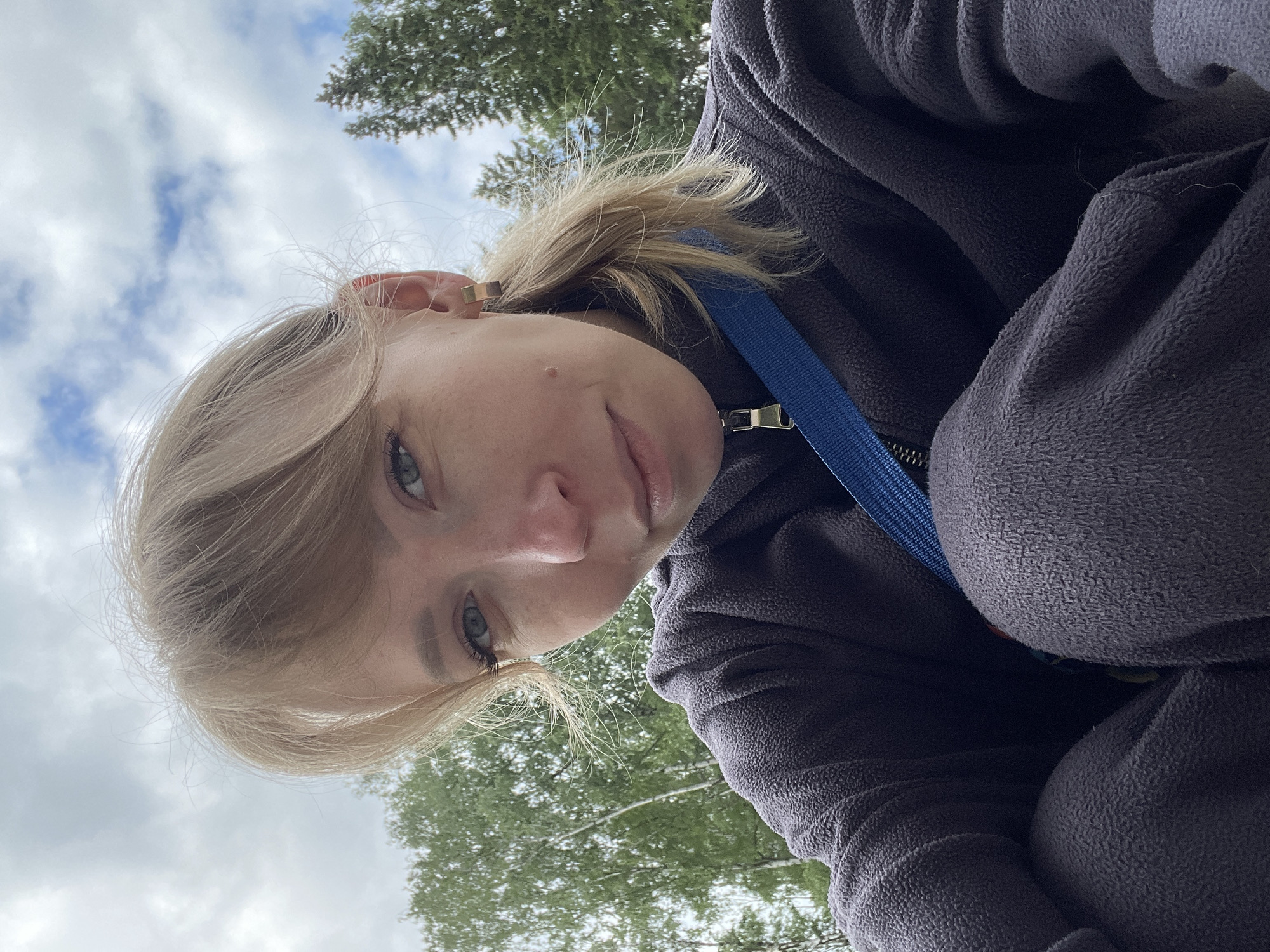 The media file is a close-up selfie of a young person, likely female, captured from a low, upward-looking angle. The subject has blonde hair and blue eyes, and is looking directly at the camera with a gentle, slightly smiling expression. A small mole is visible on their cheek, and they wear a gold-colored ear cuff or earring on their left ear.
The person is dressed in a dark purple or charcoal gray fleece jacket, with a metallic zipper pull visible at the front. A blue strap, possibly from a bag or camera, crosses their chest.
The background reveals a daytime outdoor setting with a sky featuring white, scattered clouds against patches of blue. Green tree branches with leaves are visible above and to the sides of the person, suggesting an environment like a park or a wooded area. The lighting indicates it is daytime, under overcast but not stormy conditions. No specific location cues are visible within the image to confirm the provided context of Perm, Russia, beyond it being an outdoor scene. There is no visible text in the image.