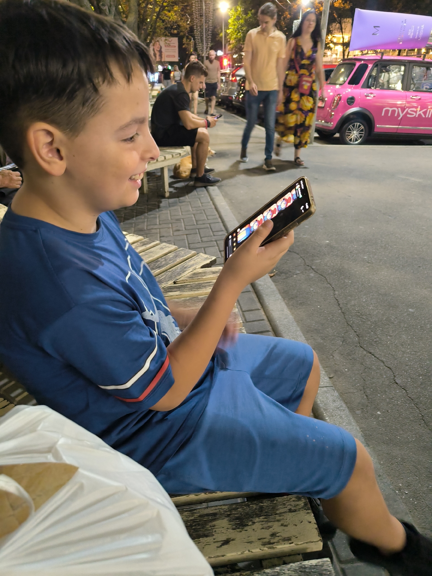 A young boy, likely under 12 years old, is seated on a wooden bench on a sidewalk in Chișinău, Moldova, engrossed in a smartphone held in his right hand. He is wearing a blue t-shirt with white and red trim on the sleeves and blue shorts. Beside him on the left is a white plastic bag. The bench appears weathered and is situated on a paved area with cobblestone detailing. In the background, several people are visible. To the boy's left, another person is partially visible, also seated on a bench. Further back, a man in a black t-shirt and shorts is seated on a bench, looking at his phone. A dog with light-colored fur lies on the ground near him. To the right of the man in black, a couple walks by; the man is wearing a light orange shirt and blue jeans, and the woman is wearing a floral dress. Behind them, a distinctive pink car with "myskin" branding and a purple promotional tube on its roof is parked on the street. The scene is illuminated by streetlights, suggesting it is evening or night. The background shows more streetlights and building facades, indicating an urban setting.