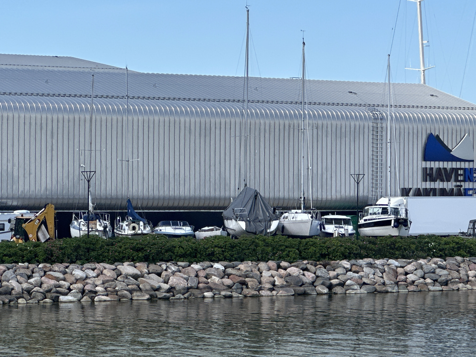 The image displays a marina facility, identified as Haven Kakumäe, located in Tallinn, Estonia. A prominent, large, modern building dominates the background, featuring a vertically corrugated metallic facade and a curved, ridged metallic roof. Masts of several sailboats extend above the roofline.
In front of the building, a row of approximately twelve boats is stored on land. These include several sailboats, some with their masts erected and wind vanes visible, and various motorboats. One sailboat is partially covered with a dark tarp. To the far left, a yellow Caterpillar excavator is parked alongside what appears to be a recreational vehicle or trailer. A white utility trailer or box truck is partially visible on the far right.
A dense, green hedge separates the boat storage area from a rock-lined shore. In the immediate foreground, a body of water exhibits light ripples, reflecting the clear blue sky overhead. A ladder is affixed to the metallic facade of the main building. A large sign on the building's right side features a blue and white stylized logo, below which the text "HAVENS" is clearly visible, with partially obscured text beneath it likely indicating "KAKUMÄE." The sky above is clear and blue. No people are visible in the scene.