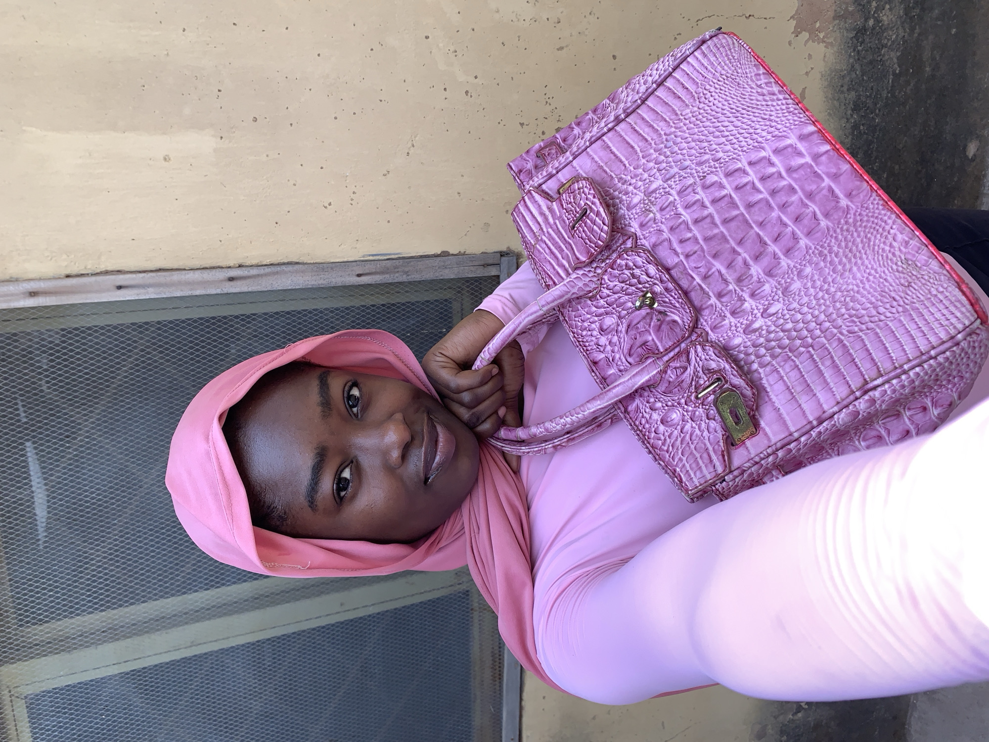 A woman with dark skin stands in Rafin Par, Nigeria, holding a pink crocodile-patterned purse. She wears a pink headscarf and a long-sleeved pink garment. The background includes a light yellow wall with a screened window.