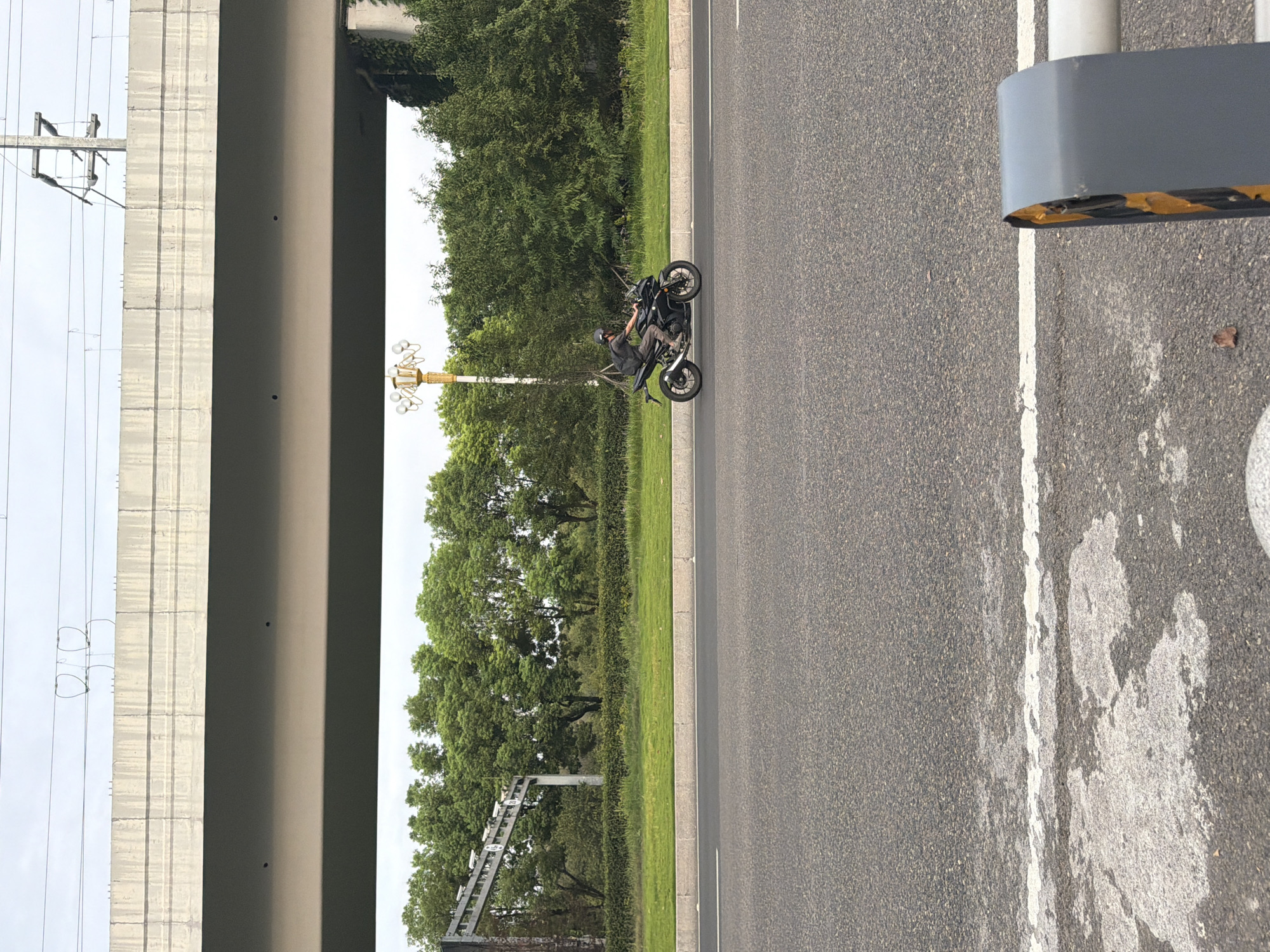 In the city of Jiangjin, China, a motorcyclist rides on a paved road beneath an elevated concrete structure. The motorcyclist, wearing a helmet and backpack, is positioned in the center-right of the frame, moving from left to right. The elevated structure, likely a bridge or overpass, dominates the upper portion of the image. Below it, a strip of green grass separates the road from a line of dense trees. A street lamp with multiple globes stands to the left of the motorcyclist. The road surface is asphalt, marked with white lines. In the foreground, a portion of a grey and yellow barrier is visible on the right side. The sky is overcast.