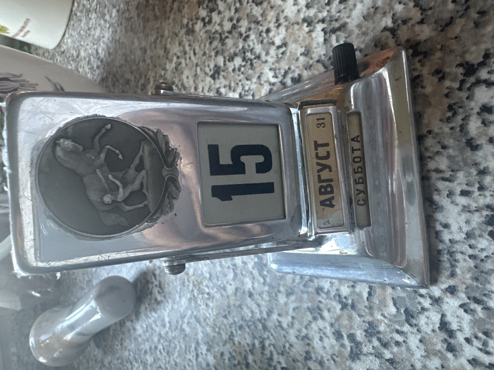 An antique metal perpetual calendar sits on a speckled countertop. The calendar is silver-toned and features a decorative circular emblem at the top depicting a person riding a horse. Below the emblem, a flip calendar displays the number "15" in large black numerals. Beneath this, another flip calendar shows the month "АВГУСТ" (August) in Cyrillic letters and the number "31". The bottom flip calendar displays "СУББОТА" (Saturday) in Cyrillic letters. A black knob for adjusting the calendar is visible on the right side. In the background, a white cylindrical object and a blurry white object resembling a handle are present. The countertop is granite with a pattern of gray and black speckles.