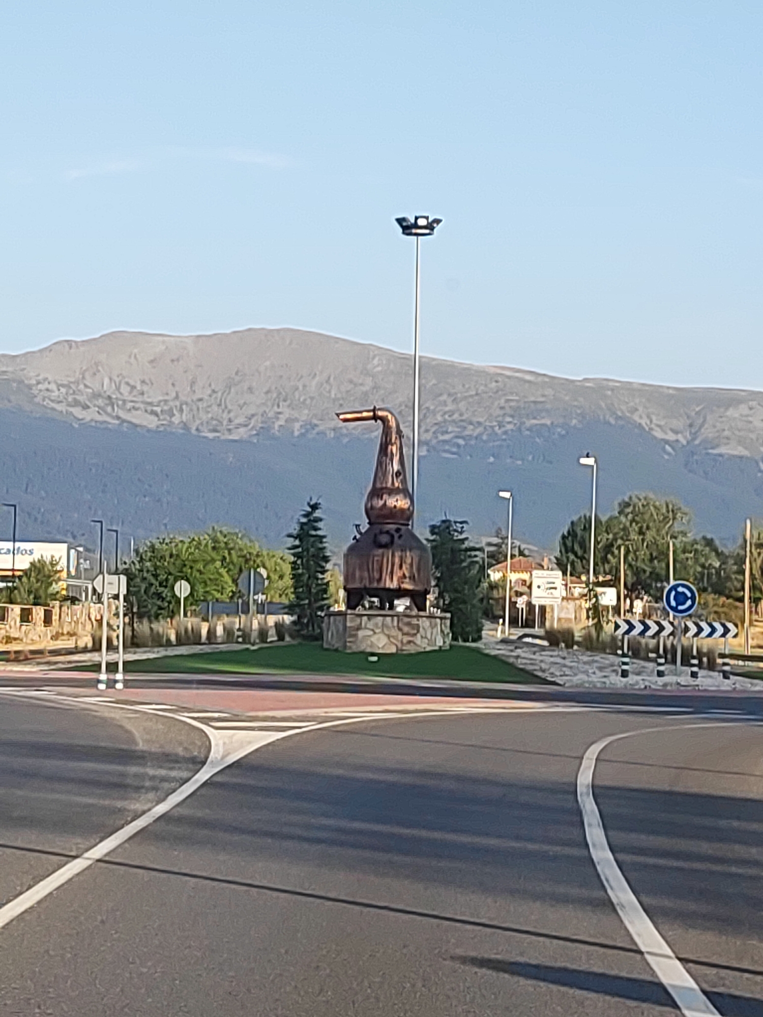 The image shows a roadside roundabout in Venta De Gabino, Spain, with a large copper sculpture of a still as its centerpiece. The sculpture, which resembles a traditional distillation apparatus with a curved spout, is positioned on a circular island surrounded by greenery and decorative stones.

In the background, a vast, rugged mountain range dominates the horizon under a clear blue sky. To the left of the roundabout, a building with signage that appears to read "Cafes" is visible, along with some smaller trees and streetlights.

The roundabout itself is paved with asphalt, marked with white lane dividers and directional arrows. Traffic signs, including a circular blue sign indicating a roundabout and chevron signs for traffic guidance, are present. The scene is illuminated by natural daylight, suggesting it was taken during the day. No people are visible in the image.