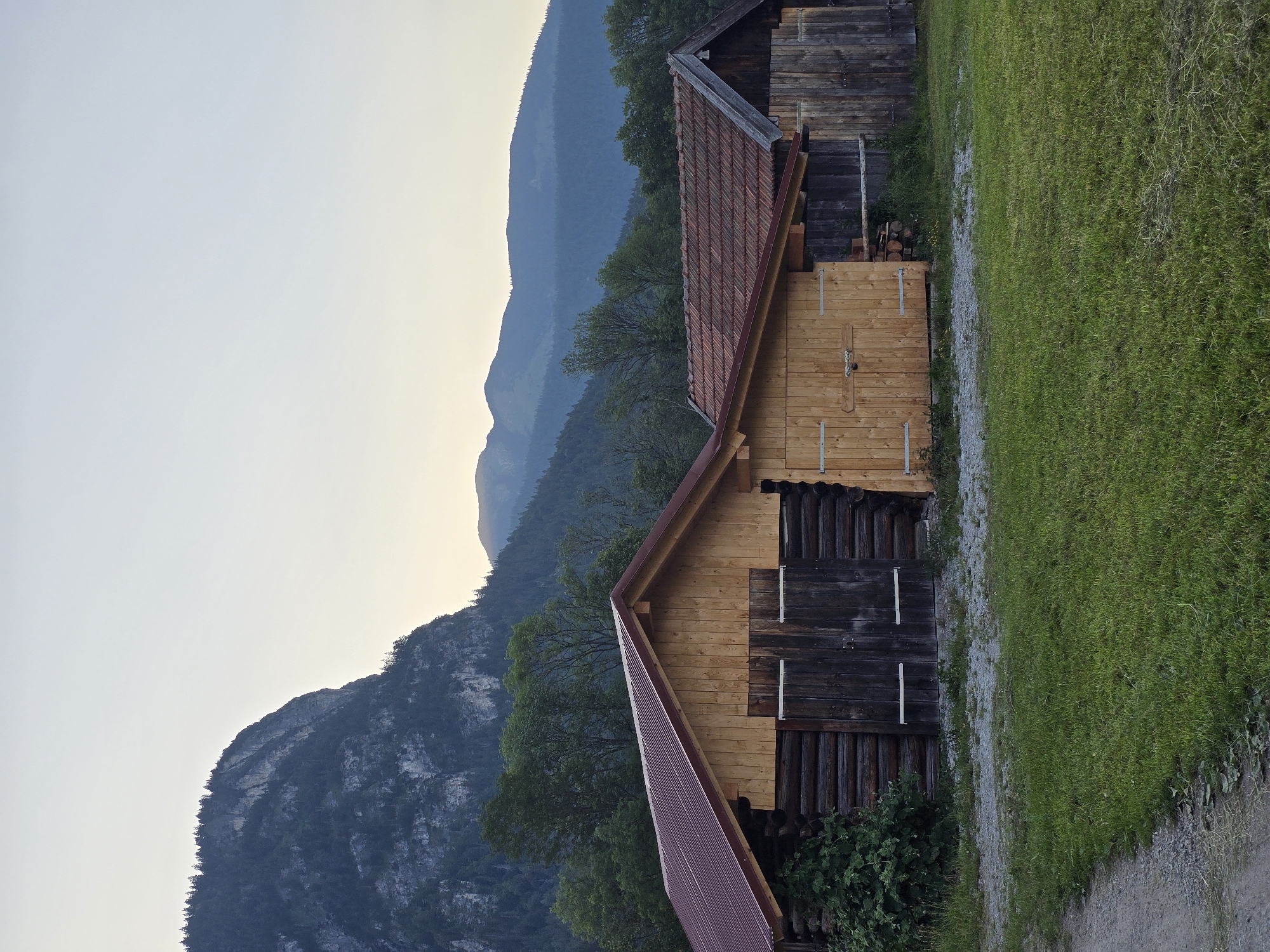 The image depicts two wooden barns situated in a grassy field with a gravel path leading to the left. In the background, a steep, rocky mountain covered with trees rises against a pale sky. Further in the distance, a range of blue, hazy mountains is visible. The barns appear to be old, with one constructed from rough-hewn logs and the other from vertical wooden planks. Both have pitched roofs, one covered in dark red corrugated material and the other with reddish-brown tiles. The scene suggests a rural or mountainous setting, likely in the Bavarian Alps, given the context of Garmisch-Partenkirchen, Germany. There are no people or animals visible in the image, and no actions or interactions are taking place. The environment is natural and tranquil, with the lighting indicative of either early morning or late evening.