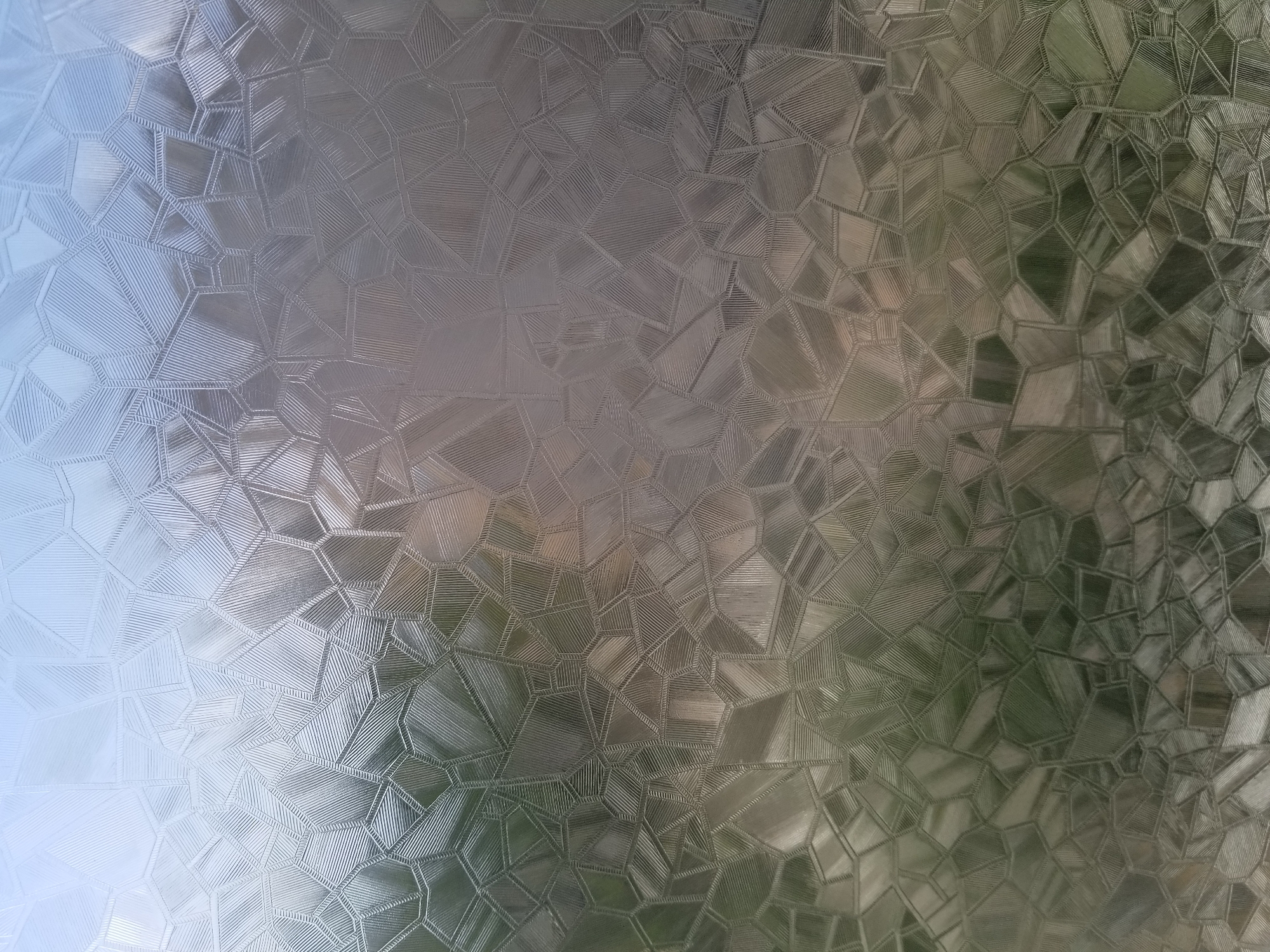 The image displays a close-up view of textured glass with a geometric pattern. The glass is translucent, allowing light to pass through, and it distorts the view of whatever is behind it, creating an abstract, dappled effect. The pattern consists of irregular polygons with fine parallel lines etched within each segment, giving the glass a frosted or embossed appearance. The colors visible through the glass are muted, suggesting an outdoor scene with greenery and possibly sky. The image does not provide any specific visual cues that would allow for the identification of a location within the city of Tiraspol, Moldova, or any other specific geographical area. There are no people, subjects, objects, or actions depicted that would indicate a particular setting or event.