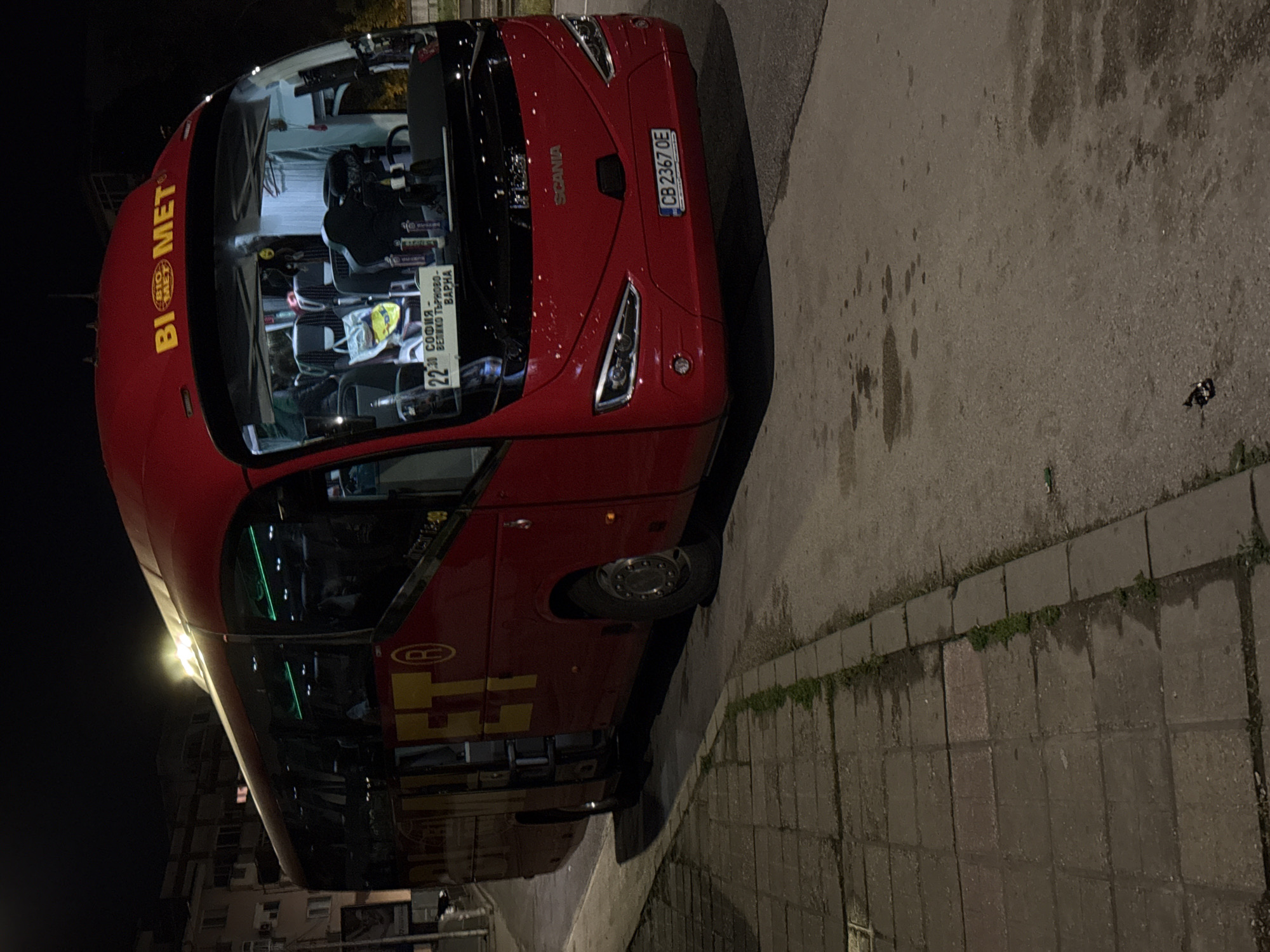 A red SCANIA bus, registration number CB 2367 OE, operated by BIOMET, is parked on a paved street in Veliko Tarnovo, Bulgaria. The bus displays a route sign indicating "22 Sofia - Veliko Tarnovo - Varna." The environment is dark, suggesting it is nighttime. The bus is positioned near a curb and a sidewalk, with a building visible in the background. No people are directly visible outside the bus.
