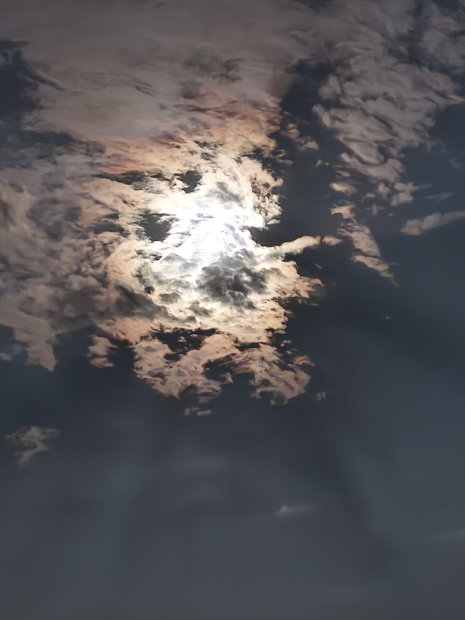 This image is a vertical, close-up shot of a sky filled with dramatic clouds illuminated by a bright light source, likely the sun, which is partially obscured. The clouds are a mix of dark, shadowed masses and lighter, brightly lit areas with a reddish-orange and white hue, suggesting a sunset or sunrise. The lighting creates a sense of depth and texture within the cloud formations. The lower portion of the image fades into a darker, more uniform sky, possibly indicating a transition to night or a heavily overcast area. There are no people, structures, or discernible landmarks visible, making it impossible to pinpoint the location within Passignano sul Trasimeno, Italy, based solely on the visual information provided. The scene is entirely natural, focusing on the atmospheric conditions of the sky.
