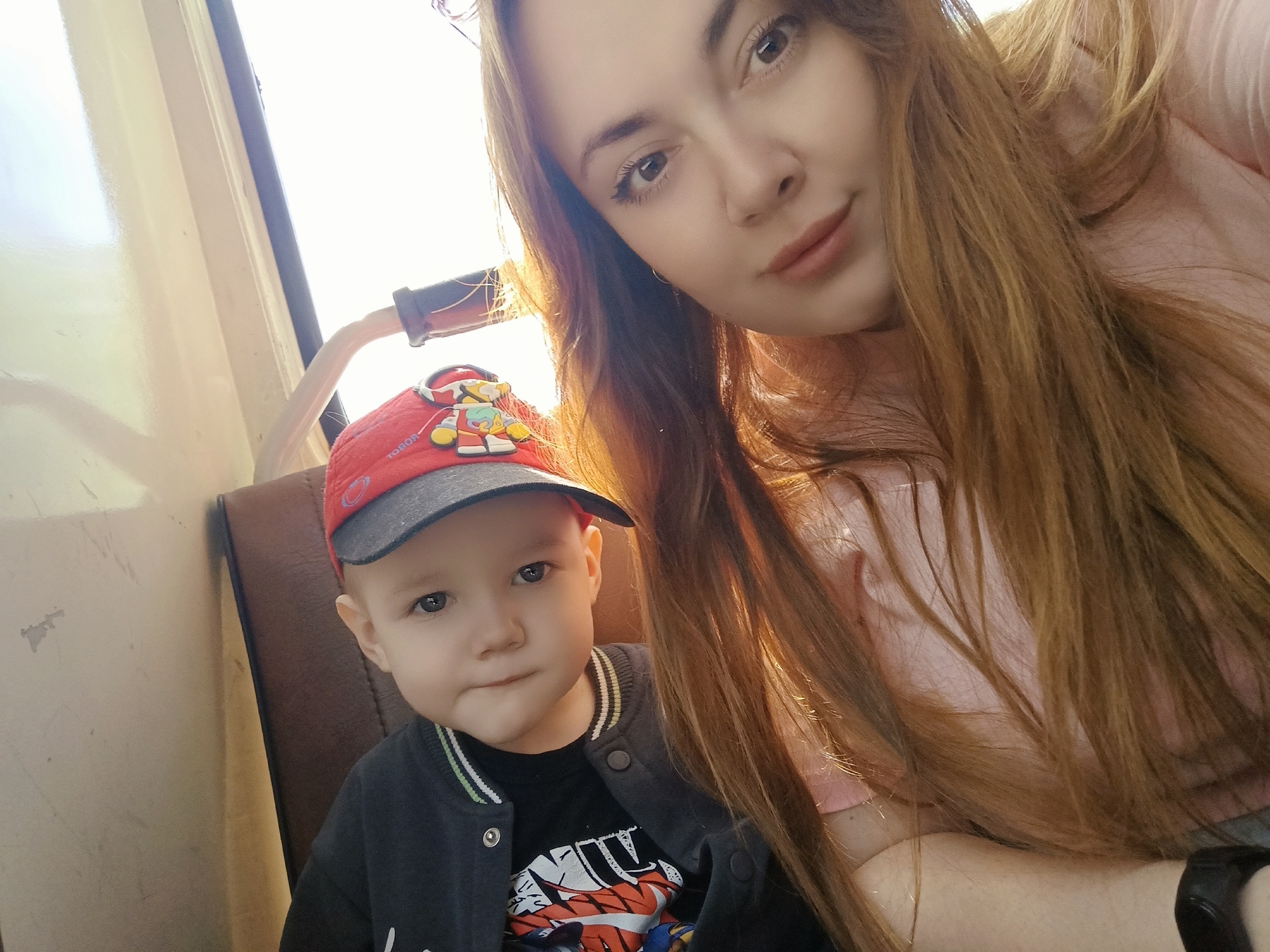 The image depicts a selfie taken indoors, likely within a vehicle, featuring a young woman and a toddler. The woman, positioned in the upper right quadrant of the frame, has long, light-brown hair and fair skin. She wears a light-pink top. The toddler, located in the lower-left quadrant, wears a red and grey baseball cap and a black jacket over a printed t-shirt. The setting is identifiable as an interior space, possibly a bus or train, due to the presence of a window and a seat. There is no information to directly confirm this image was taken in Chaikovsky, Russia.