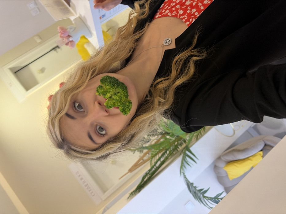 A blonde female is captured in a brightly lit indoor setting, humorously posing with a large piece of broccoli held between her lips, which are obscured. Her eyes are wide, creating an exaggerated, surprised expression. She wears a black blazer over a red dress adorned with white floral patterns, and a silver necklace with a 'Tree of Life' pendant is visible around her neck. Her left hand is partially visible on the right, holding a mobile device.
The background reveals a modern reception or waiting area. To the right, a white counter holds a white ceramic vase containing pink, clustered decorative elements. Behind the counter, a yellow cushion or object is visible. A white door with a silver 'WC' sign, indicating a restroom, is present in the mid-right. To the left, a large, leafy green indoor plant stands next to a beige, tufted armchair with a bright yellow cushion. Above another white door on the left, a sign reads "COSMETOLOG," indicating the establishment is a cosmetology studio or clinic. This scene is located within a cosmetology studio or clinic in the city of Budva, Montenegro.