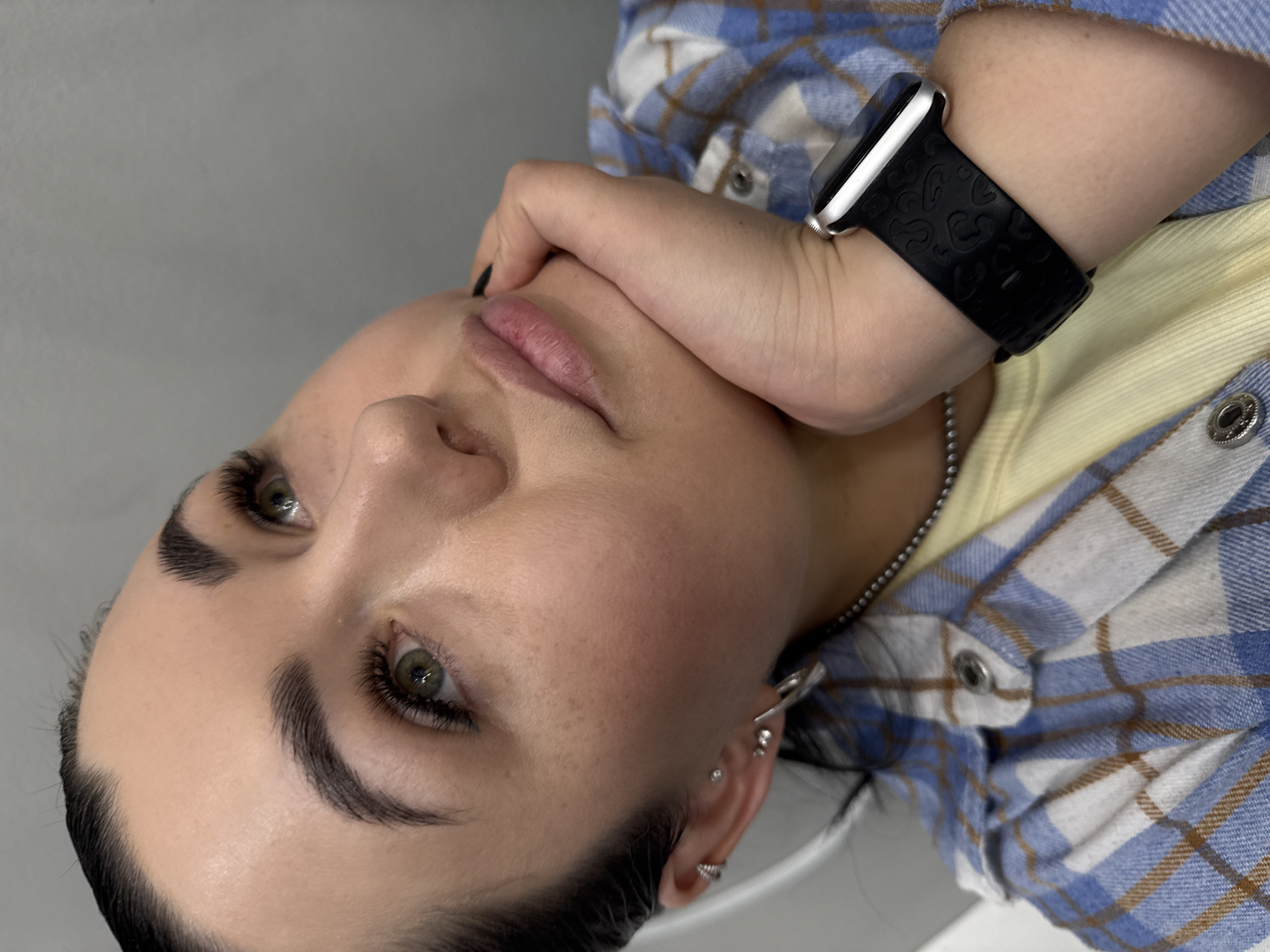 A close-up, slightly rotated portrait features a young adult indoors. The individual has fair skin, dark hair, prominent dark eyebrows, and green eyes, with long, dark eyelashes. Their lips appear subtly tinted. The subject's right hand, with black painted fingernails, rests against their chin and jawline.
They are dressed in a light-colored undershirt, possibly yellow, beneath a blue, white, and brown plaid flannel shirt with silver snap buttons. A thin silver-colored chain is visible around their neck. On their left wrist, they wear a silver-cased smartwatch with a black, patterned silicone band. Their right ear displays multiple piercings, including a helix, tragus, and a textured hoop in the lobe.
The background is a plain, light gray or off-white wall, indicating an indoor setting. The lighting is soft and even. The individual is looking slightly upwards and to the left. No visible text is present. The general aesthetic suggests a focus on beauty or personal style, potentially related to beauty services given the enhanced facial features. The image's origin is Chaikovsky, Russia, though no explicit location cues are visible within the frame.
